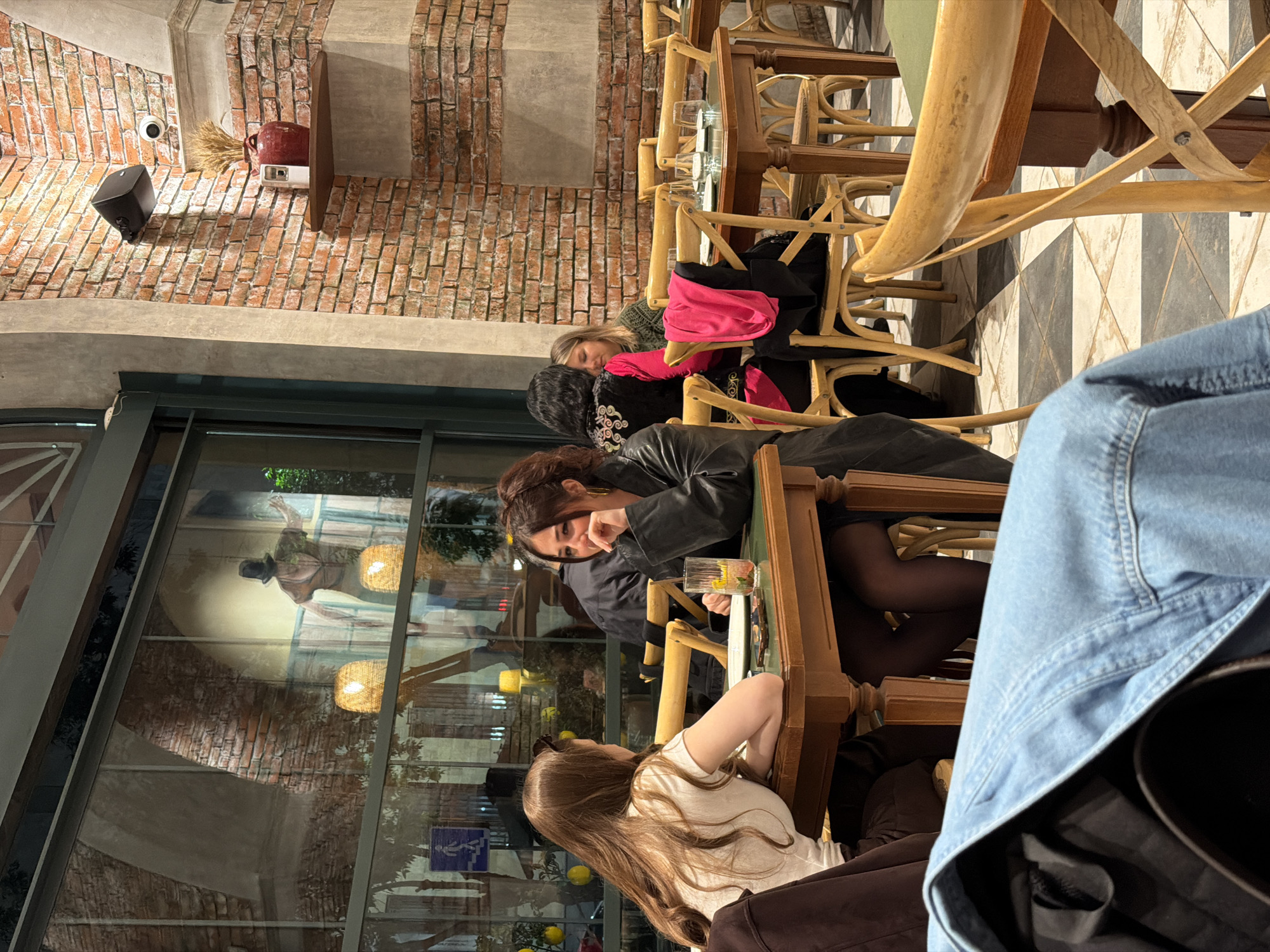 The image captures a scene at a restaurant in Yerevan, Armenia, where three women are seated at a wooden table. The woman closest to the camera has long, light brown hair and is wearing a white shirt. Another woman in a black leather jacket is seated next to her, holding a glass with a beverage inside. A third woman, further away from the camera, is seated at the end of the table and wears an olive green sweater with a pink jacket.
The restaurant has a rustic interior with exposed brick walls and large windows that reflect an outdoor setting. The seating arrangement includes light-colored wooden chairs, and the floor has a black and white checkered pattern. A surveillance camera, a speaker, and a decoration are visible on the brick wall.