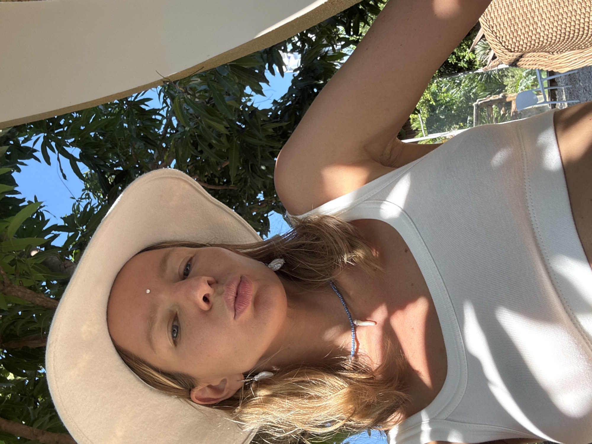 A close-up, overhead, selfie-style shot shows a blonde woman wearing a wide-brimmed, light-colored hat and a white tank top. She has a single, small, clear stud in her forehead between her eyebrows. She is wearing a blue beaded necklace with a shell pendant and shell earrings. Her lips are slightly parted. She is outdoors, with green foliage and a bright blue sky visible in the background. The lighting suggests it is daytime, likely sunny, with dappled shadows on her upper body and the tank top. The setting appears to be tropical, with lush green leaves and trees. A woven basket is visible in the upper right corner of the frame, partially obscured. The overall mood is relaxed and summery. No text is visible.