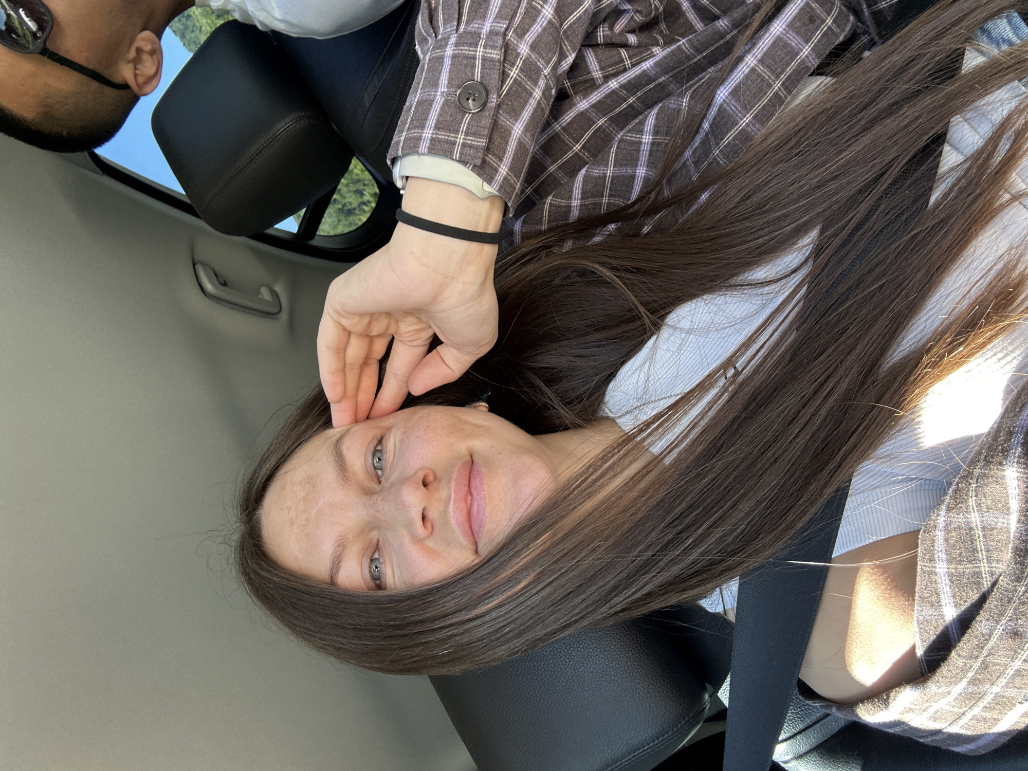 A young woman with long, dark brown hair and blue eyes smiles into the camera, her right hand resting near her temple. She is seated in the back seat of a car, indicated by a black seat and seatbelt visible in the frame. She wears a white top and a brown and purple plaid jacket. Partially visible in the upper right corner is the head of another person, wearing sunglasses and a white shirt, also in the car. The background outside the car window suggests a rural or natural setting with green foliage and a bright blue sky. The lighting indicates it is daytime, possibly sunny. The image appears to be a selfie taken inside a vehicle, likely on a road trip. The metadata does not specify a location within Jabuka, Montenegro, but the environment visible is consistent with a rural or mountainous region.