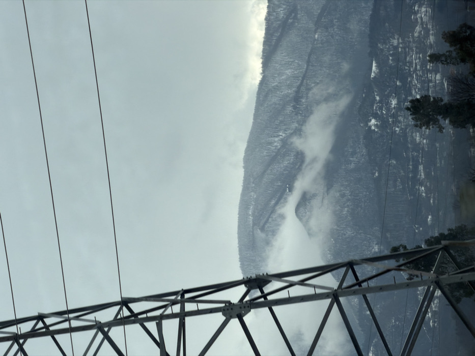 The image captures a winter scene in Terjola, Georgia. The left foreground features a section of a galvanized steel lattice transmission tower, with multiple taut, dark-colored power lines extending horizontally across the upper frame. Snow-covered peaks and slopes dominate the background, densely vegetated with trees exhibiting snow accumulation. Low-altitude atmospheric moisture, consistent with fog or clouds, partially obscures the mid-section of the mountains. A cluster of residential structures, characterized by dark rooftops against the snow, is visible on the lower slopes and valley floor, indicative of the city of Terjola. Deciduous trees with bare branches are present in the lower right foreground. The sky is uniformly overcast and light grey.