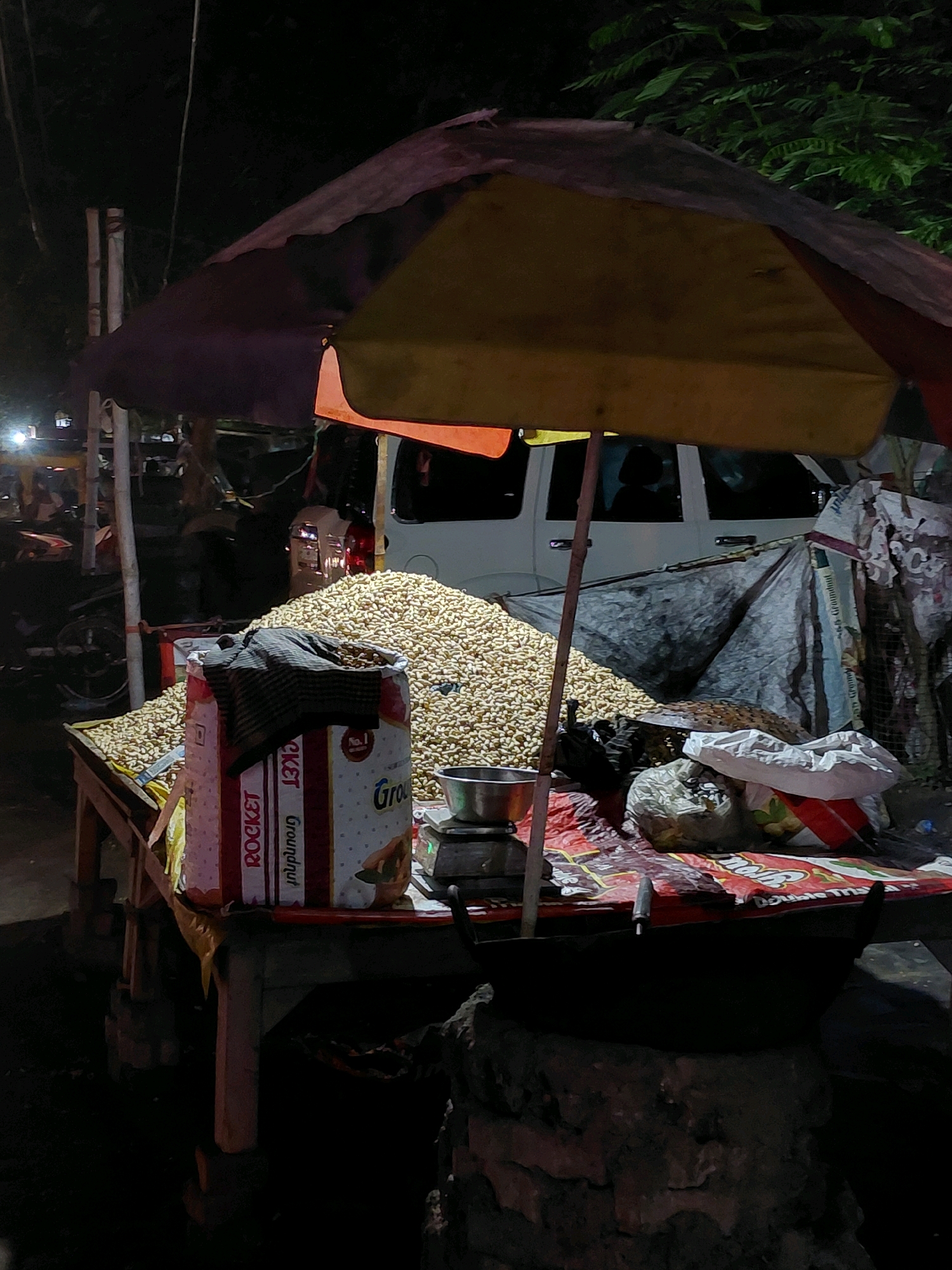 A street vendor's stall, likely in Patna, India, is depicted at night. A large pile of peanuts sits on a wooden table, covered in part by a multi-colored umbrella that is partially unfurled, revealing yellow and red hues. To the left of the peanut pile, a large sack labeled "ROCKET Groundnut" is visible, with a striped cloth draped over its opening. A small weighing scale is placed on the table next to the peanuts. In front of the table, a black wok rests on a brick structure, suggesting food preparation. The background shows dimly lit trees and a white vehicle, indicating an outdoor urban setting. The scene is illuminated by artificial lights, creating a somewhat grainy and dark atmosphere.