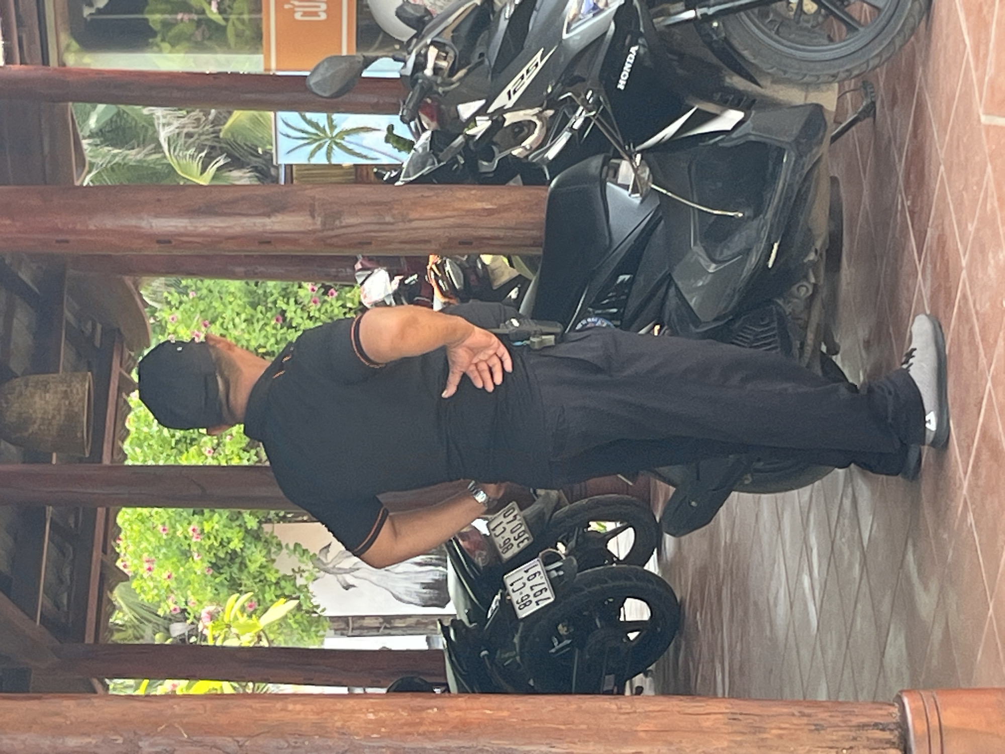 A man wearing a black cap and black polo shirt with orange trim stands beside several motorcycles under a covered, open-air structure. The man has his left hand on his hip and his right hand resting on his thigh. He is wearing dark pants and light gray shoes. Two motorcycles are visible, one black with a license plate reading "86-C1 797.91" and another black with a license plate reading "86-C1 36040". Behind the man and motorcycles, lush green foliage with pink flowers is visible. A bell-like object hangs from the ceiling of the structure. The floor is tiled.