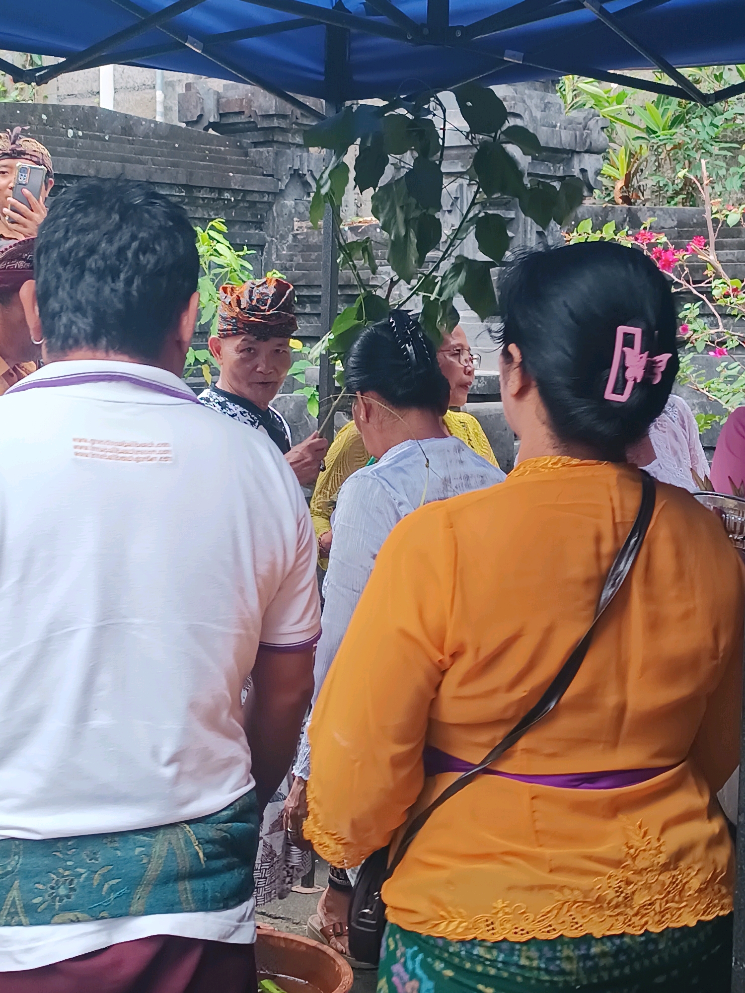 The image depicts a gathering under a blue tent, likely for a cultural or religious event in Tabanan, Indonesia. In the foreground, the back of a person wearing a white polo shirt with purple trim is visible. They are also wearing a sarong with a green and floral pattern. To their right, another person, seen from the back, wears an orange blouse with a purple sash and intricate embroidery on the hem, also with a green and patterned sarong. Their dark hair is held back with a pink butterfly clip.
Further into the scene, a man in a traditional Balinese headdress and a patterned shirt is looking towards the foreground. Behind him, a woman in a yellow garment and glasses is visible. Another individual, partially obscured, is holding a smartphone to take a picture. The background features traditional Balinese stone architecture and lush greenery, including flowering plants. The overall atmosphere suggests a community event or ceremony taking place outdoors, possibly within a temple complex or a traditional compound.