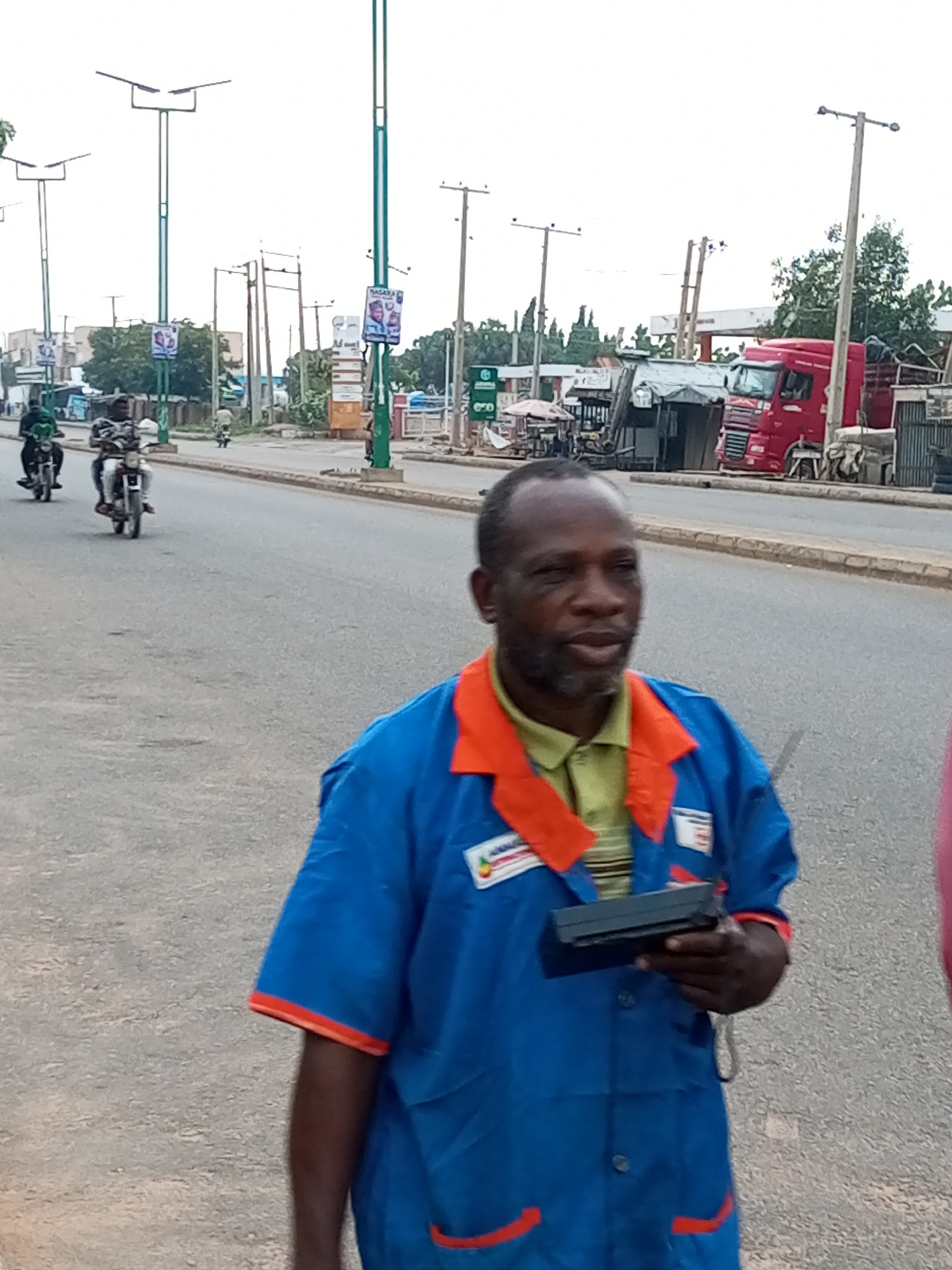 In Garko, Nigeria, a man wearing a blue uniform with orange trim and a green collared shirt underneath is standing on a paved road. He has dark skin and short, graying hair. He is holding a dark rectangular object, possibly a radio or scanner, in his right hand. The road is lined with streetlights and poles. In the background, several motorcycles are visible on the road, with riders. There are also buildings and what appears to be a red truck in the distance. The sky is overcast.