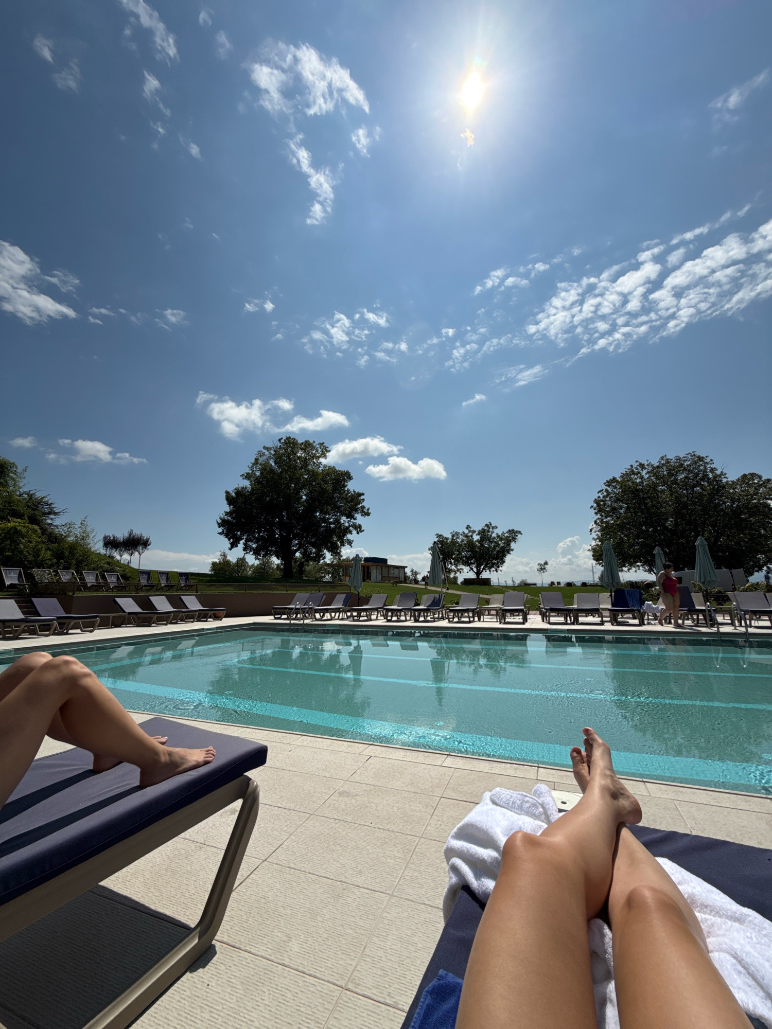 The image depicts a sun-drenched day at a resort swimming pool, potentially located in Kvareli, Georgia, given the context. In the foreground, two individuals are relaxing on lounge chairs, their legs prominently visible. One person on the left reclines on a dark blue lounger with their legs extended towards the pool. Another person on the right is also on a dark blue lounger, with a white towel draped over it, and their legs are positioned similarly.
The swimming pool itself is a focal point, with clear blue water reflecting the bright sky. A blue border is visible along the edge of the pool. Surrounding the pool are numerous empty lounge chairs and umbrellas, suggesting a resort or hotel setting.
In the background, a lush green landscape unfolds. Large, mature trees are scattered across the terrain, with one particularly prominent, expansive tree dominating the mid-ground on the right. Beyond the immediate pool area, a gentle slope rises, covered in vegetation. A few low-lying buildings or structures are visible nestled amongst the trees in the distance. The sky is a vibrant blue, adorned with scattered white clouds and a bright, slightly diffused sun positioned in the upper center of the frame. The overall atmosphere is one of relaxation and leisure on a warm, sunny day.