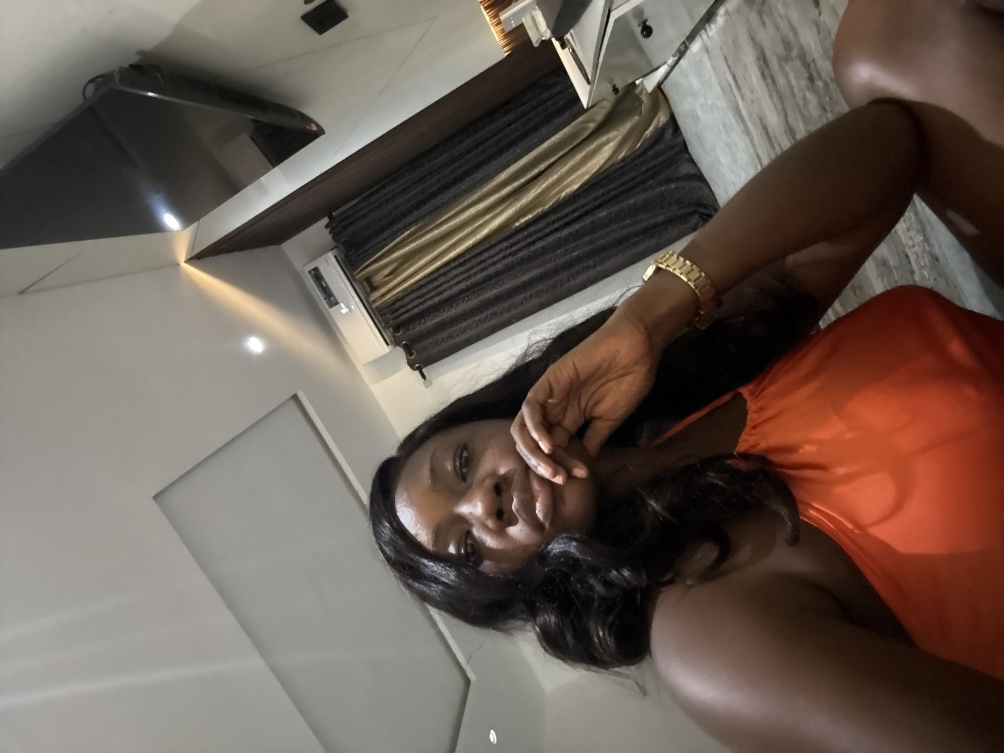 A woman is visible from the chest up, wearing an orange garment and a gold-colored bracelet on her left wrist. She is in an interior space with light-colored walls and marble flooring. A television is mounted high on the wall. Behind her are gray and gold curtains. There is no definitive evidence to verify the specific location in Iddo Tudunwada, Nigeria.