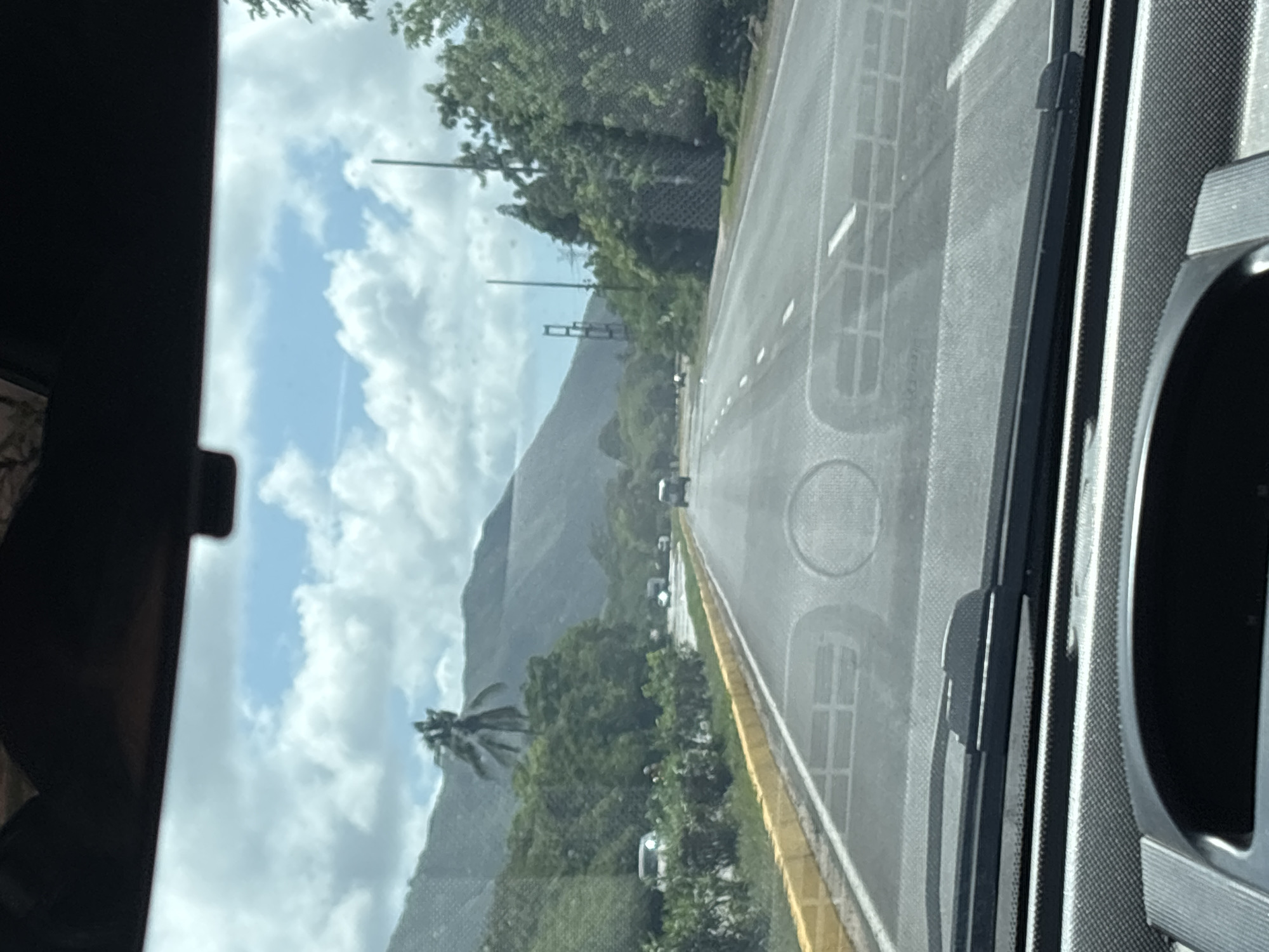 This image captures a view from inside a vehicle, looking out through the windshield at a highway scene. The road ahead is paved and has clear white lane markings. On the left side of the road, there is a yellow curb with a white line running parallel to it. Beyond the curb, there are green bushes and trees, and in the background, a lush green mountain range under a cloudy sky. Several vehicles are visible on the highway, including a light-colored car on the left shoulder and other cars further ahead. Power lines and poles are present along the right side of the road, interspersed with dense green foliage. The dashboard of the vehicle, including the windshield wipers and part of the steering wheel, are visible in the foreground. The overall scene depicts a highway journey through a scenic, possibly tropical, environment.