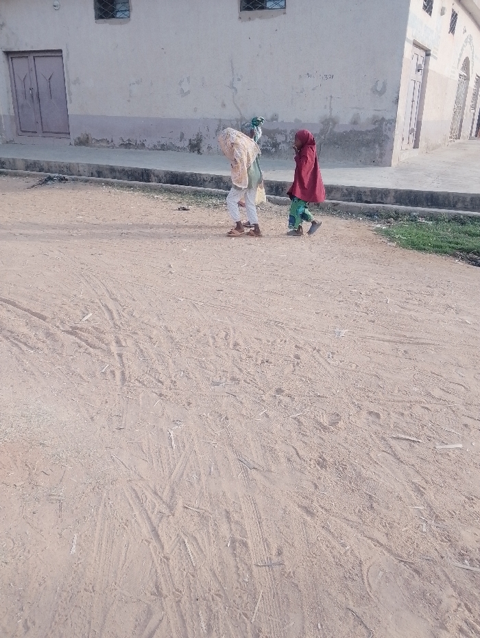 Two individuals, likely children, walk along a dirt road beside a light-colored building. The person on the left is mostly covered by a light tan shawl, while the person on the right wears a red headscarf and a knee-length red coat. The road shows signs of vehicular traffic, with tire tracks visible in the dirt. The building has a light-colored facade with a few windows and doors. A small patch of green vegetation is visible to the right of the road.
