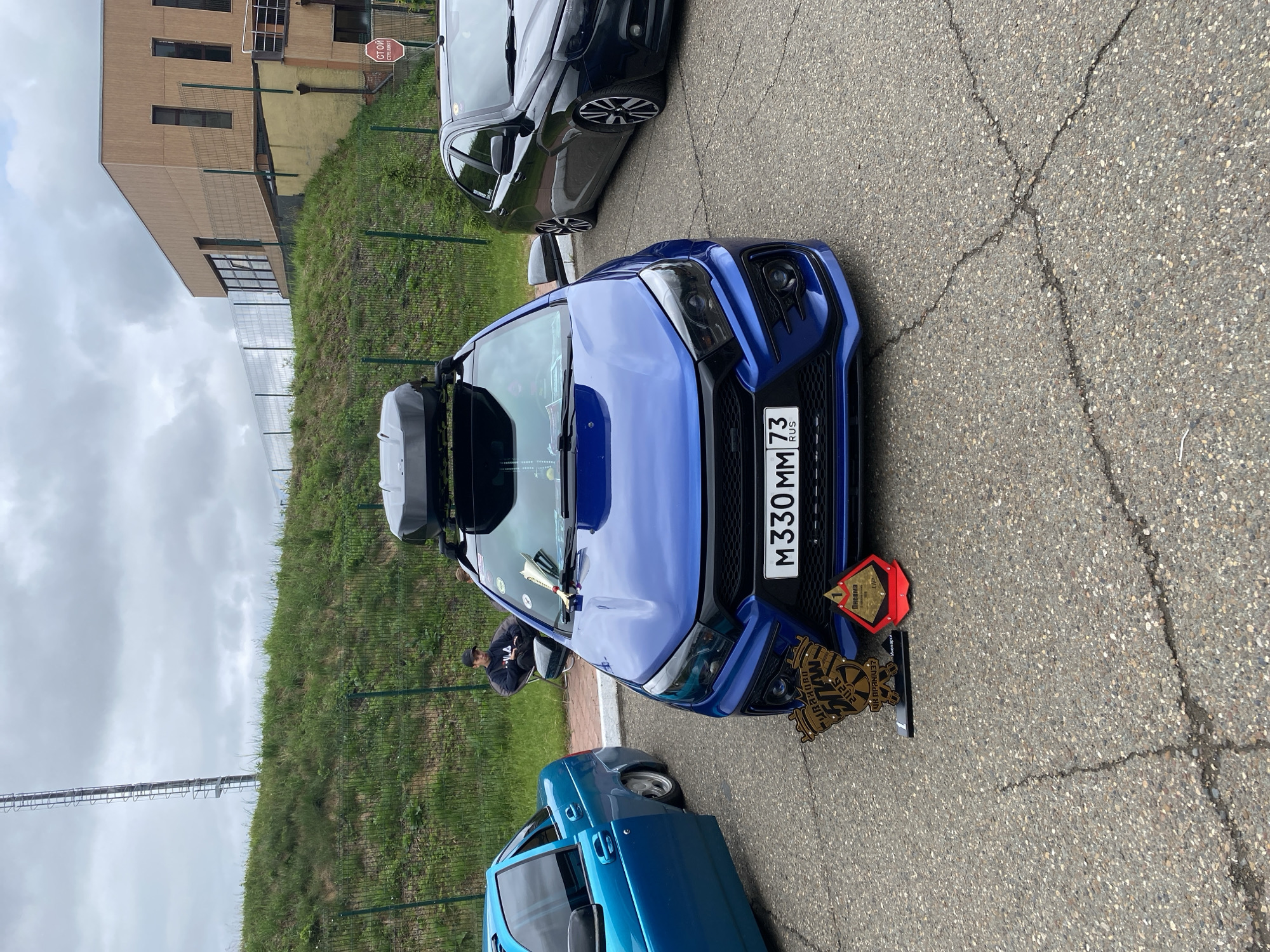 The image captures an outdoor car event in Прикамский, Russia, under an overcast sky. Three modified sedans are parked on an asphalt surface with visible cracks.
In the foreground and center, a royal blue sedan, likely a Lada Vesta, is prominently displayed. It features a custom black grille, blacked-out headlights, a sporty front bumper, and a roof-mounted black cargo box. The front license plate reads "M330MM 73 RUS." Two awards are placed on the ground directly in front of this vehicle: a large, light brown trophy resembling wood, inscribed with Cyrillic text including "Иваново 2026," and a smaller red, triangular trophy with the number "1" and the Cyrillic word "Пневма" (Pneuma/Air) along with "LIP." A white, cone-shaped object is visible on the car's dashboard through the windshield.
To the immediate left of the central blue car, a light blue sedan with significantly lowered suspension is partially visible, with its front passenger door slightly ajar. To the right, a black sedan, also featuring lowered suspension and black and silver multi-spoke wheels, is partially visible.
In the background, behind a green chain-link fence, a grassy hill slopes upward. A single individual is seated in a dark, collapsible chair on a red-paved area at the base of the hill. This person is wearing a dark hoodie, possibly with a white logo, and a dark baseball cap. Further back, on top of the hill to the left, a tall, metal lattice tower stands against the cloudy sky. To the right, a modern building with light brown paneling and dark-framed windows is situated above the hill. A red circular "СТОЙ" (STOP) sign with additional Cyrillic text is visible near the building.