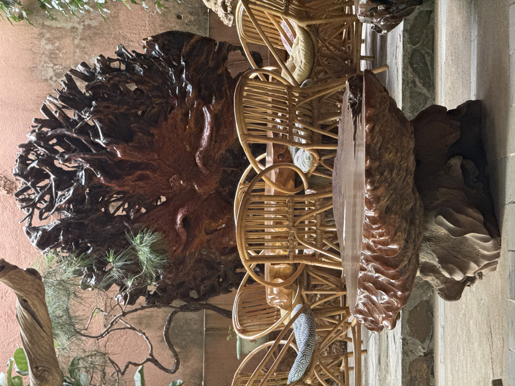 The scene captures a partially obscured view of what appears to be an outdoor or semi-outdoor seating area with artistic wooden installations, likely in Bản Đêu, Vietnam, given the prompt. Five rattan chairs are stacked and slightly rotated, occupying the center. An elaborate, dark brown, root-like wooden sculpture occupies the left side, with the texture suggesting complex natural forms. A light brown, rectangular wooden piece is on the right. The background consists of a neutral-colored wall and stone detailing, offering a contrast to the textures of the wooden installations.