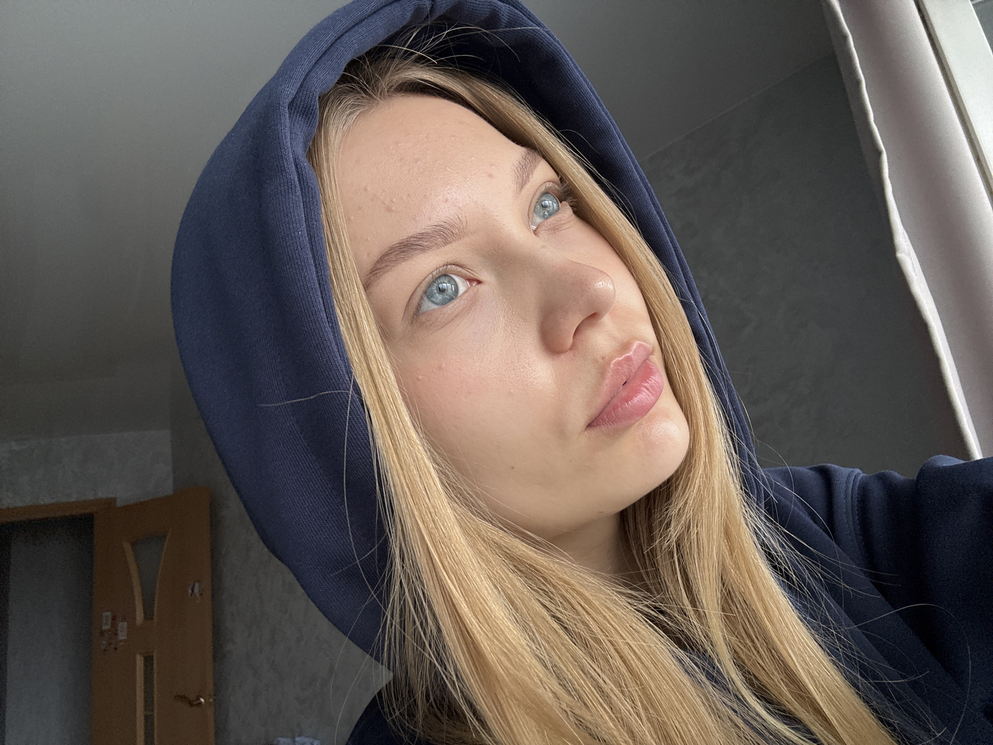 A young woman with light blonde hair and blue eyes is wearing a dark blue hoodie with the hood up. She is looking upward and to the right. Her facial expression is pensive. The background is blurred, but a window with a light-colored curtain and a wooden door are partially visible. The photograph was taken in what appears to be a room in a residential building in Chaikovsky, Russia.
