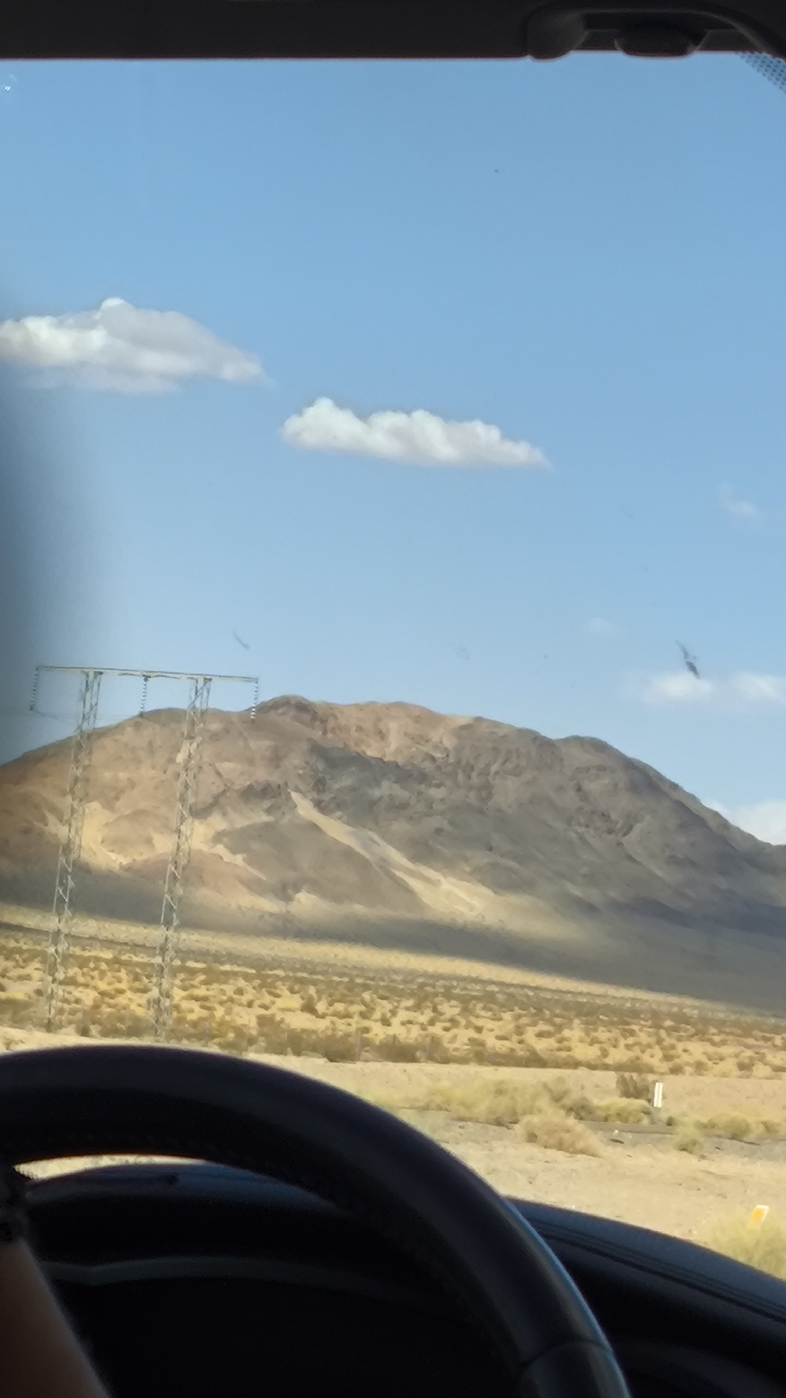 A vertical, medium shot captures a desert landscape through a car's windshield. The foreground is dominated by the black, curved steering wheel of a car, partially obscuring the view. Beyond the wheel, a vast expanse of dry, brush-covered desert stretches towards a large, tan-colored mountain range in the background. The mountain is illuminated by sunlight, highlighting its rugged texture and varied coloration. A tall electrical transmission tower with metal lattice supports stands to the left of the frame, in front of the mountains. The sky above is a clear, bright blue with a few scattered white clouds. The scene is consistent with a view from a vehicle traveling through the desert region of Newberry Springs, California, United States.