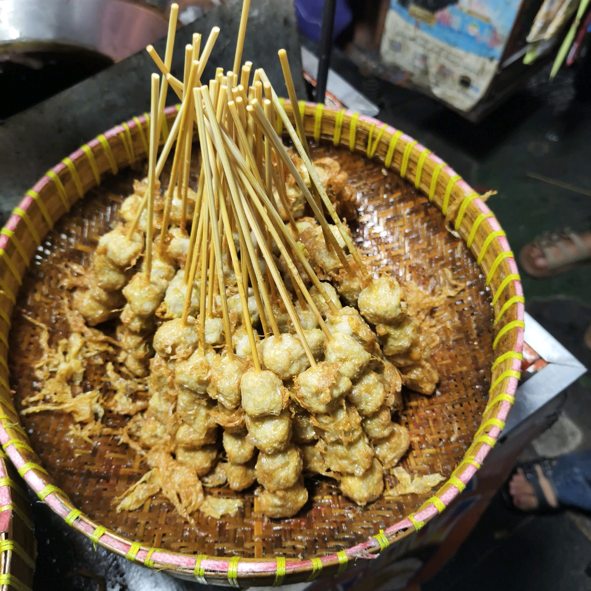 In the city of West Jakarta, Indonesia, a close-up, eye-level shot captures a woven bamboo basket overflowing with fried meatballs on skewers. The basket itself is intricately woven, with a decorative rim adorned with yellow and pink bands. The meatballs are golden brown, with a slightly crispy texture, and are skewered with thin wooden sticks. The background is blurred, but hints at a bustling street food environment, with other vendors and customers visible. The lighting is dim, typical of evening street market conditions, with a warm glow illuminating the food.