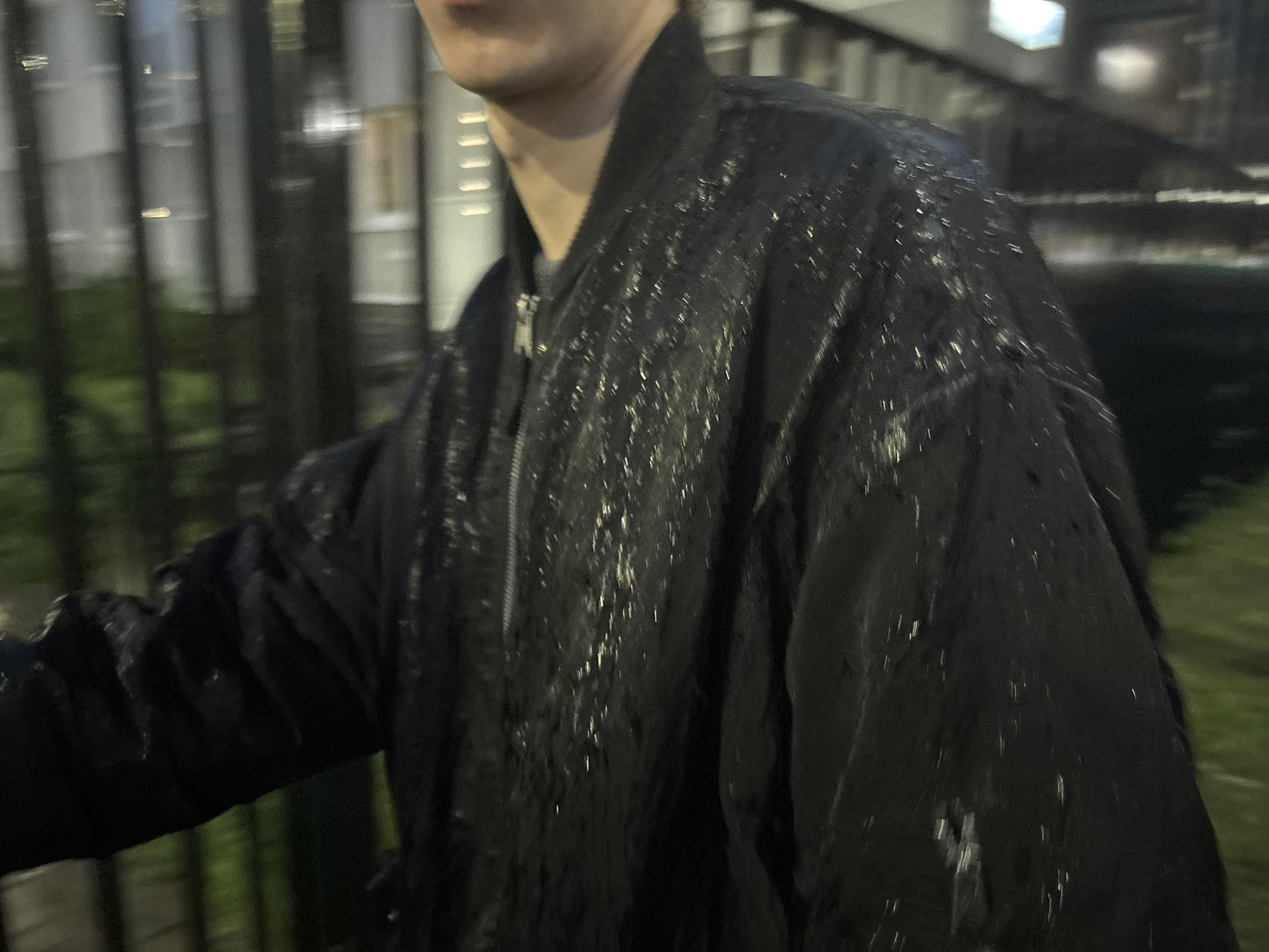 The image is a close-up, low-angle, outdoor shot of a person wearing a black bomber jacket that appears to be wet, with water droplets or streaks visible on the surface. The jacket has a zipper down the front. The background is blurred and appears to be an urban environment at night, with vertical lines suggesting fencing or railings, and green areas that could be grass or trees. Lights from buildings or streetlights are also visible in the background. The motion blur suggests movement.