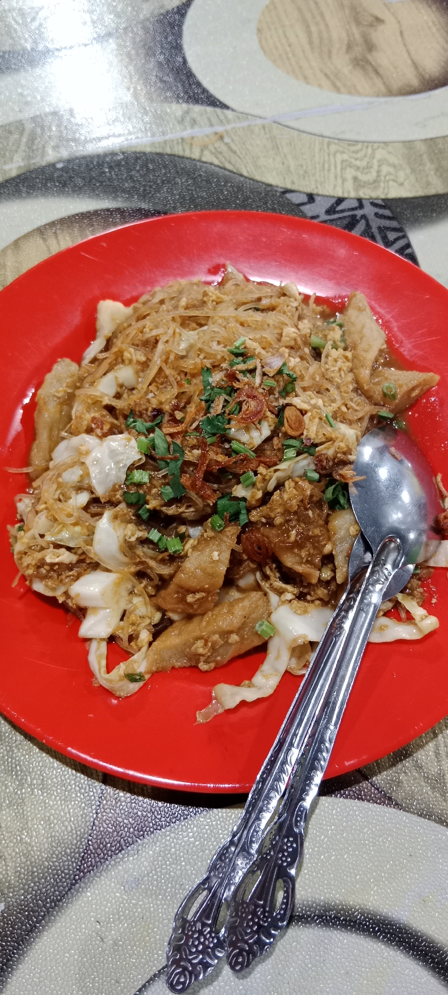 A close-up, top-down shot displays a portion of a red plate filled with a dish featuring translucent noodles, sliced white cabbage, and pieces of fried food, all coated in a savory sauce. The dish is garnished with chopped green onions and crispy fried shallots. A silver spoon rests on the right side of the plate, partially submerged in the food. The plate is set on a patterned tablecloth with beige, brown, and gray tones. The overall impression is of a restaurant setting, likely in Tanjung Pinang, Indonesia, given the commonality of such dishes in Indonesian cuisine.