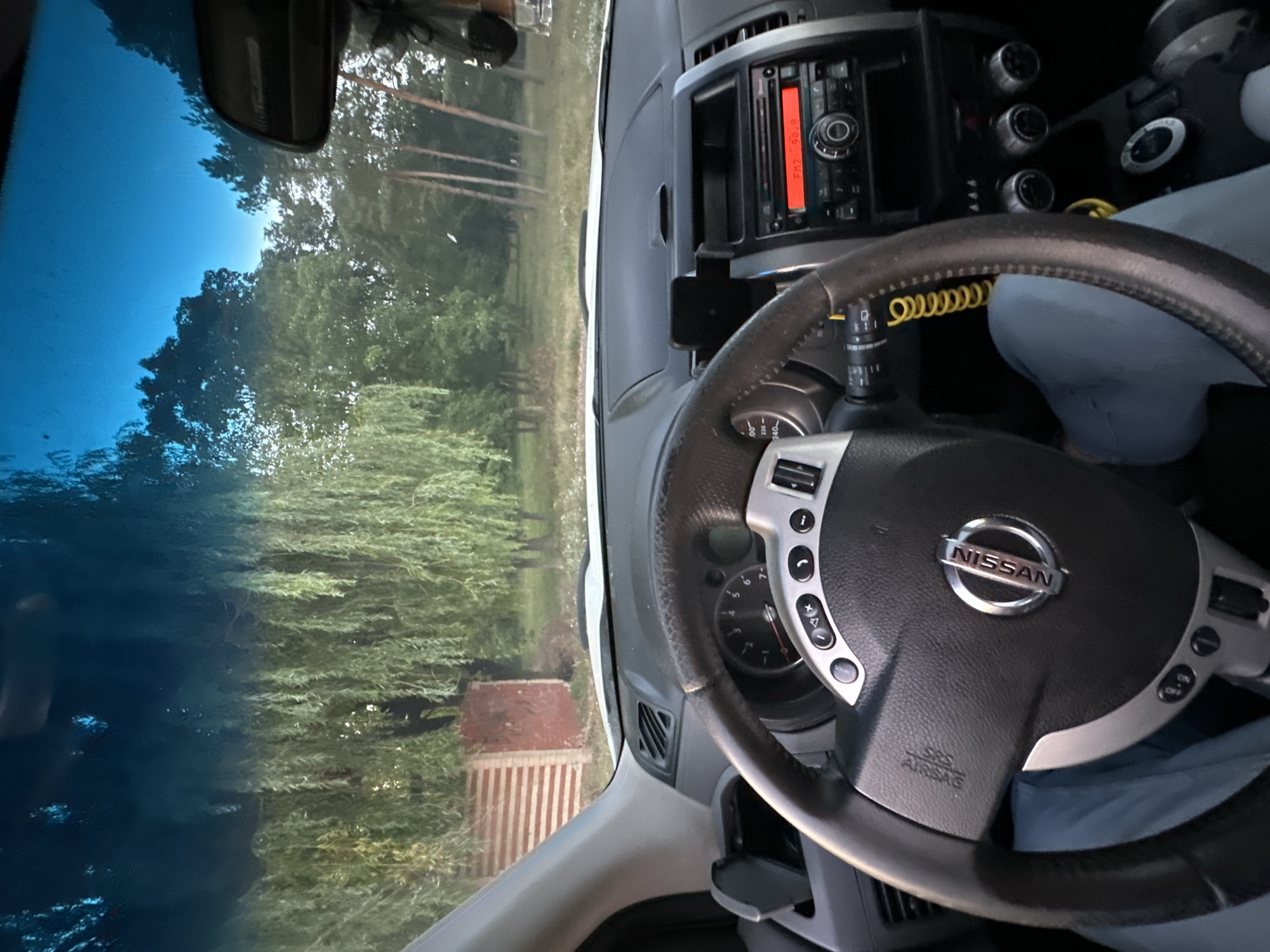 The image shows the interior of a car, likely a Nissan, viewed from the driver's perspective. The steering wheel, dashboard with a radio display, and a portion of the driver's legs wearing light blue jeans are visible. Through the windshield, a rural landscape with trees and a building with striped siding is seen. The radio display shows "FM2 98.0". The overall scene suggests the car is parked in a natural setting, possibly on the outskirts of Makiv, Ukraine, given the context.