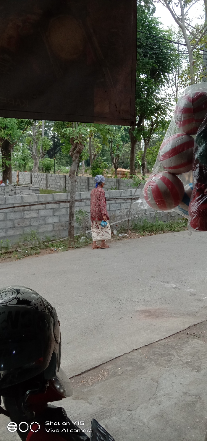 The image depicts a street scene in Temenggungan, Indonesia, captured from a low vantage point, likely from within a roadside stall or vehicle. In the immediate foreground, a black motorcycle helmet with faded white markings is visible on the lower left, attached to the front of a red and black scooter or motorcycle. The ground consists of a light grey paved surface (asphalt or concrete) exhibiting a central seam and minor debris. To the upper right foreground, several white and red striped balls, likely volleyballs, are visible, encased in clear plastic bags and suspended from an unseen fixture, suggesting merchandise for sale.
A single elderly individual walks on the paved surface in the midground, moving away from the viewer towards the left. The person is dressed in a patterned, long-sleeved, reddish-brown top, a light-colored long skirt or sarong with a darker pattern, and light-brown flip-flops. A blue head covering is worn, and a small, light-blue pouch is carried in the right hand. Behind this individual, a low retaining wall constructed of light grey concrete blocks extends horizontally across the scene, forming a series of partitioned sections. A slender, gnarled tree trunk rises between the paved path and the concrete wall.
The background is dominated by numerous mature trees with green foliage and light-colored trunks, indicating a verdant, possibly rural or park-like setting. The upper portion of the image is obscured by a dark, weathered banner or awning, displaying faint, inverted lettering "KAREL" on its lower left section, along with indistinct oval patterns. The scene is illuminated by natural daylight.