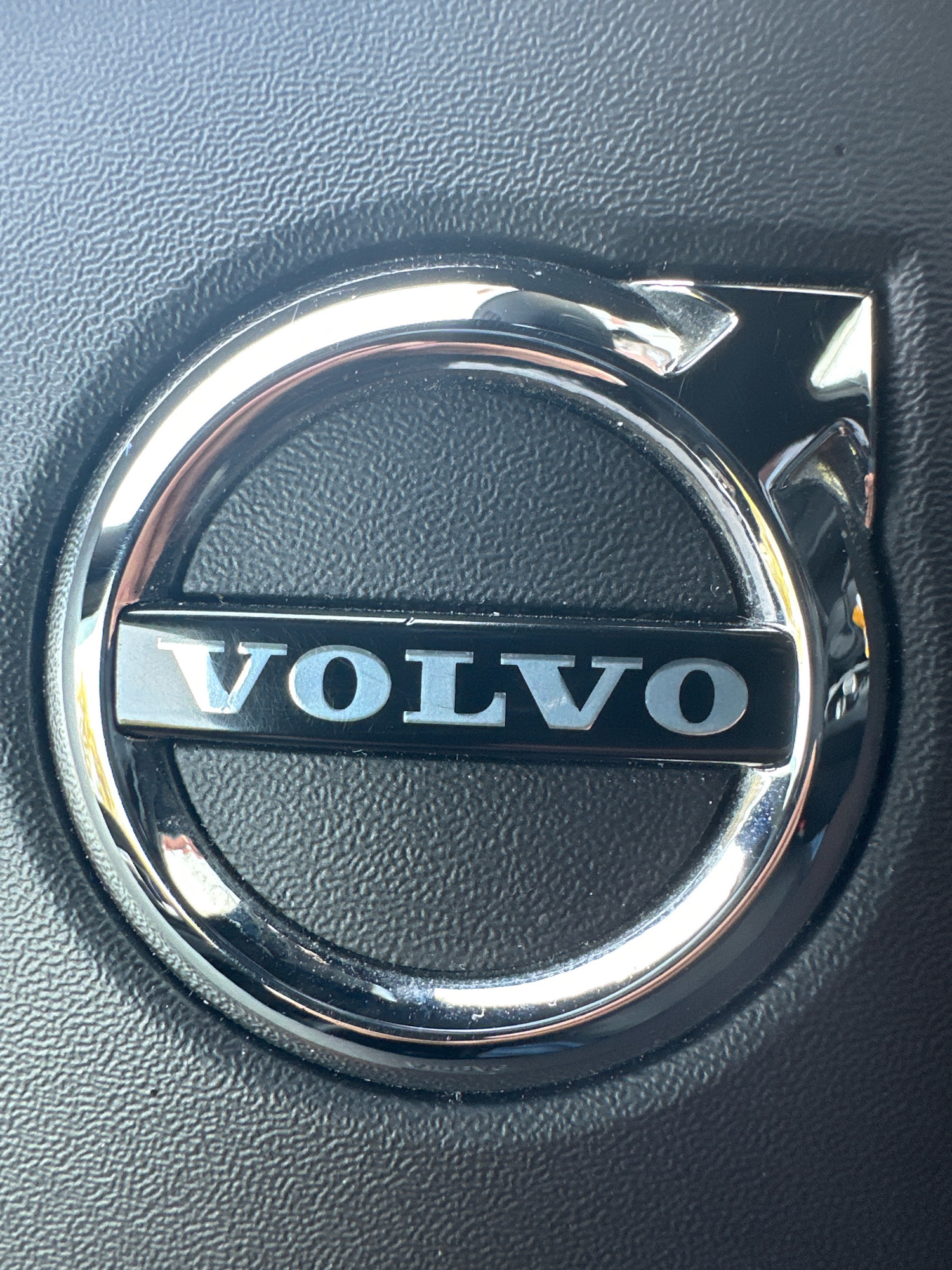 A close-up shot focuses on the silver Volvo logo emblem, centrally positioned on a black, textured surface, likely part of a car's interior. The emblem features the word "VOLVO" in silver, sans-serif capital letters against a black rectangular background. A metallic silver ring surrounds this rectangle, and the overall emblem is slightly raised from the textured surface. The lighting suggests an indoor environment with reflections visible on the polished metal of the logo. The image does not contain any information to determine its location in Murmansk, Russia, or any other specific geographic location. There are no people or actions depicted in the image.