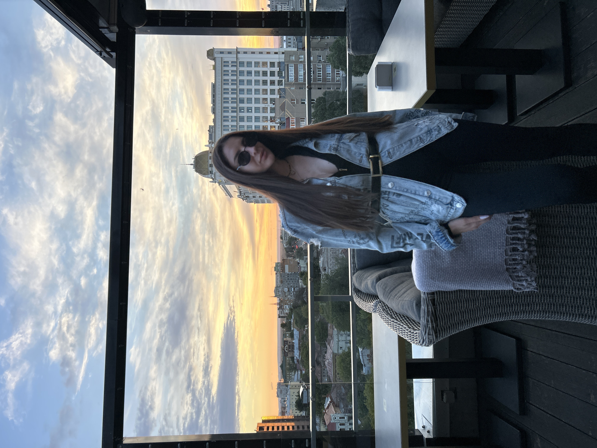A woman stands on a rooftop terrace in Novokuybyshevsk, Russia, with a cityscape and sunset in the background. She is wearing a denim jacket, black pants, and sunglasses. Tables and chairs are visible on the terrace, suggesting it's an outdoor dining or lounge area. The sky is filled with clouds illuminated by the setting sun, casting a warm glow over the urban landscape.