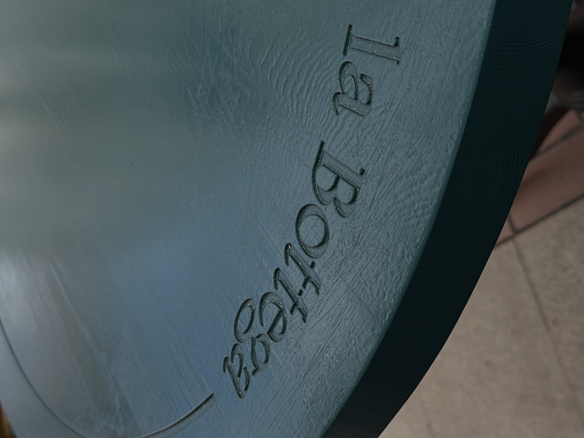 The image shows a close-up of a dark teal-colored object with embossed text. The texture of the object appears to be a rough, slightly wrinkled material, possibly leather or a textured plastic. The embossed lettering reads "la Bottega" in a stylized font. The letters are slightly raised and appear to be filled with a darker pigment or shadow, giving them a dimensional quality.
The lighting in the image suggests it might be indoors or in a shaded outdoor area, with a soft, diffused light that highlights the texture of the object and the embossed text. There are some subtle reflections on the surface, indicating a smooth, albeit textured, finish. The background is out of focus, showing hints of a beige and brown tiled surface, suggesting an indoor setting like a shop floor or a display area.
No people are visible in the image. The activity taking place is not evident beyond the presentation of the object with its branding. The overall impression is one of a product detail shot, focusing on the quality and branding of the item. Given the location context of Ufa, Russia, this object could be part of a luxury good, furniture, or a decorative item. The text "la Bottega," which translates to "the shop" or "the workshop" in Italian, suggests an artisanal or high-end brand.