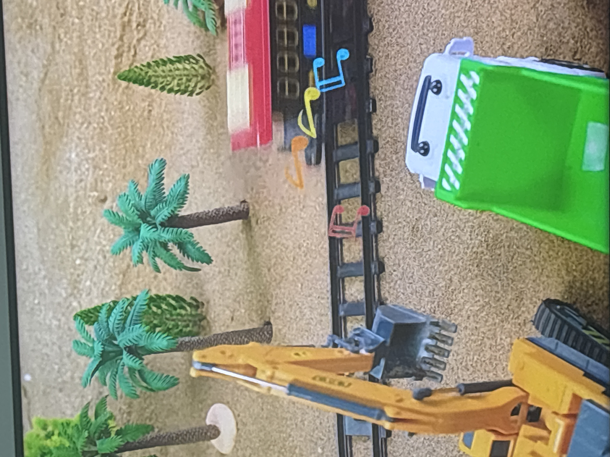 The image depicts a miniature diorama with a desert-like sand landscape. Several toy palm trees and a smaller, cone-shaped green tree are scattered across the scene. In the foreground, a yellow toy excavator with a grey bucket is positioned next to a toy train track. A toy train, with a red and white top and black body, is on the tracks. Music notes, in orange, yellow, and blue, appear to be floating above the train. Further in the foreground, a green and white toy garbage truck is visible. The scene is indoors, with a gray border at the top, suggesting it's a captured image of a display. There is no indication of this scene being located in Dubai, United Arab Emirates.