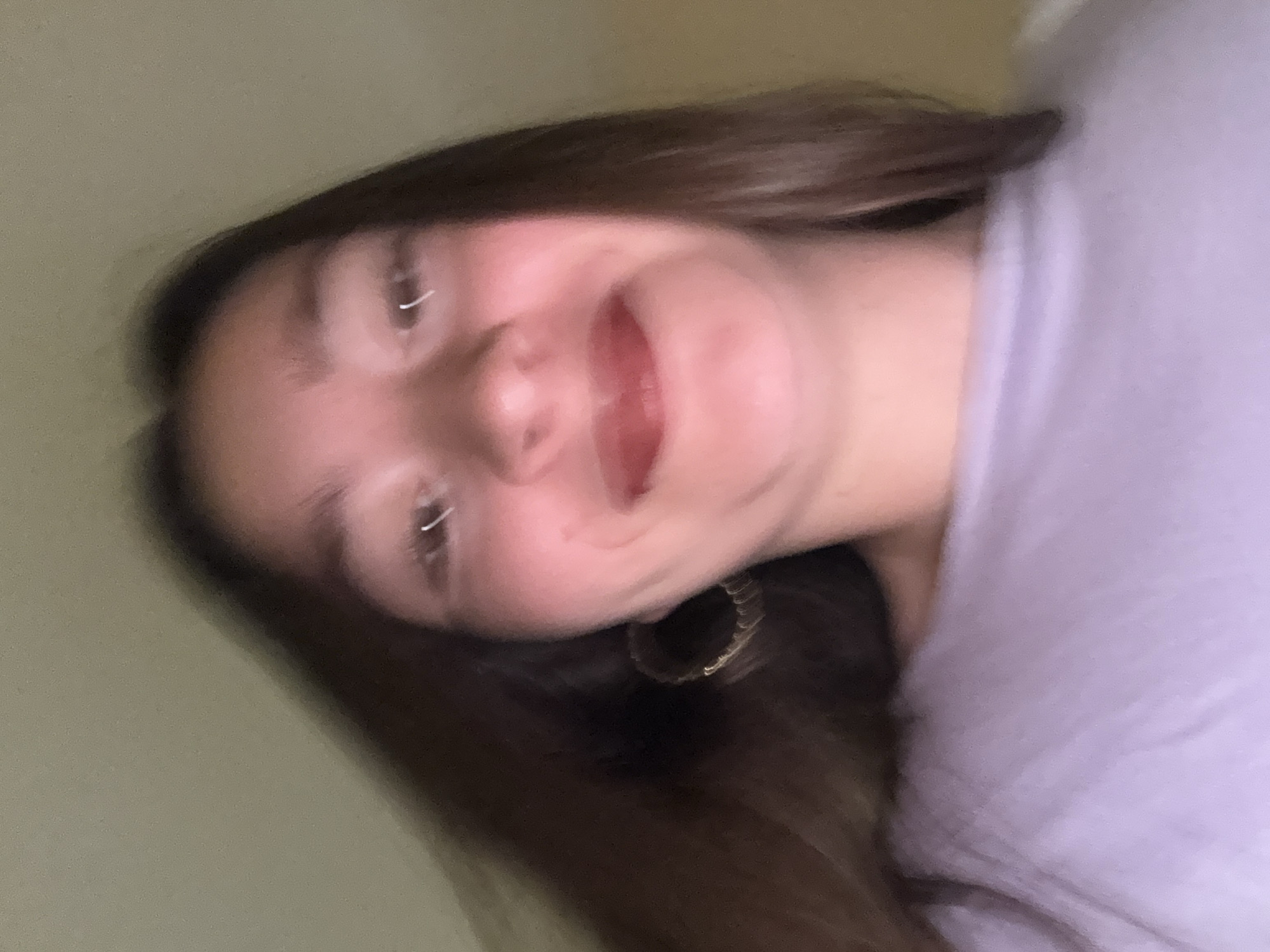 A close-up, slightly blurry, low-angle selfie shows the upper body of a young female with long, dark brown hair. She is wearing a light purple, crew-neck top. She has a noticeable hoop earring in her left ear. Her lips are colored with a pinkish-red lipstick. The background is a plain, light-colored wall. There are no identifying landmarks or context to suggest a specific location in Sofia, Bulgaria.