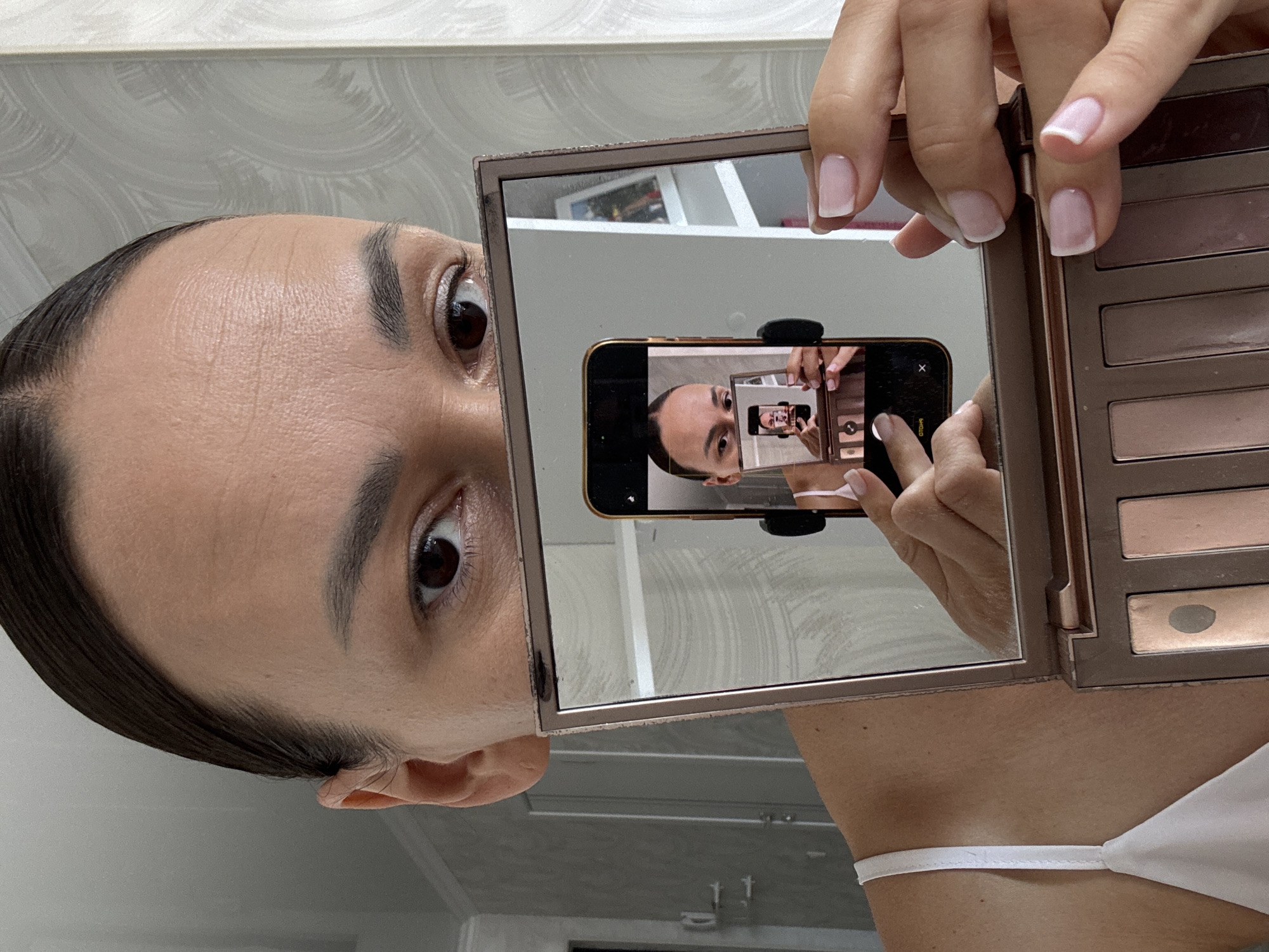 The image shows a close-up view of a woman's face as she holds up a makeup compact mirror. Her dark hair is pulled back tightly. She is looking into the compact mirror, which reflects her face and a smartphone that she is holding. The smartphone, in turn, is reflecting the compact mirror and the woman. The makeup compact is open, revealing several eyeshadow shades. The woman's fingernails are manicured with a French tip. The background appears to be an indoor setting with textured wallpaper. There is no visible indication of the city of Sochi, Russia.