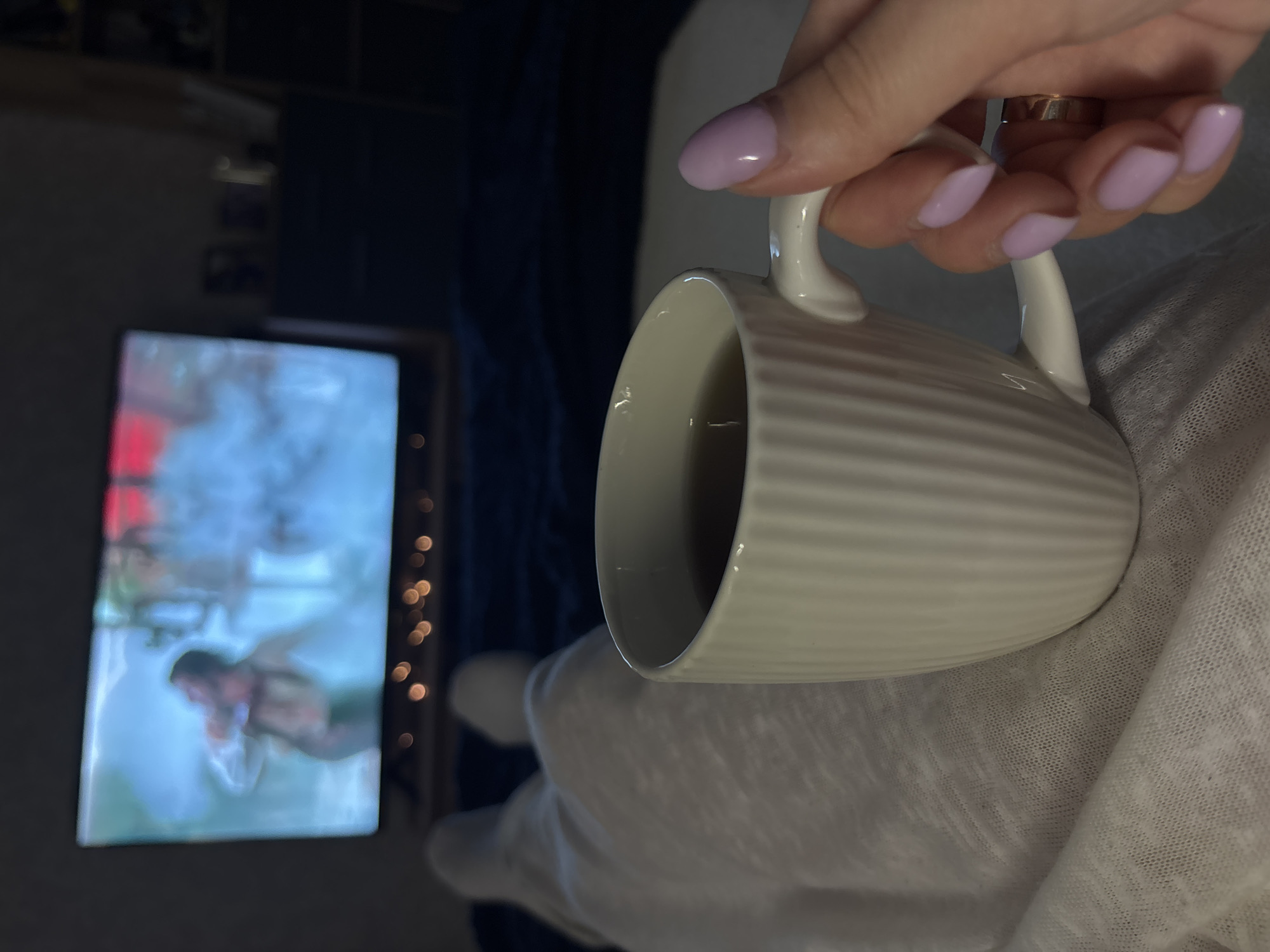 A hand with lavender-colored fingernails holds a white, ribbed mug filled with a dark liquid, possibly coffee or tea. The mug rests against a white, loosely woven fabric, likely a blanket or a piece of clothing. In the background, out of focus, is a television screen displaying a blurry image of a man. The scene is indoors, with a dark blue curtain and a string of lights partially visible behind the television. The location within the city of Chaikovsky, Russia, cannot be determined from the provided image.