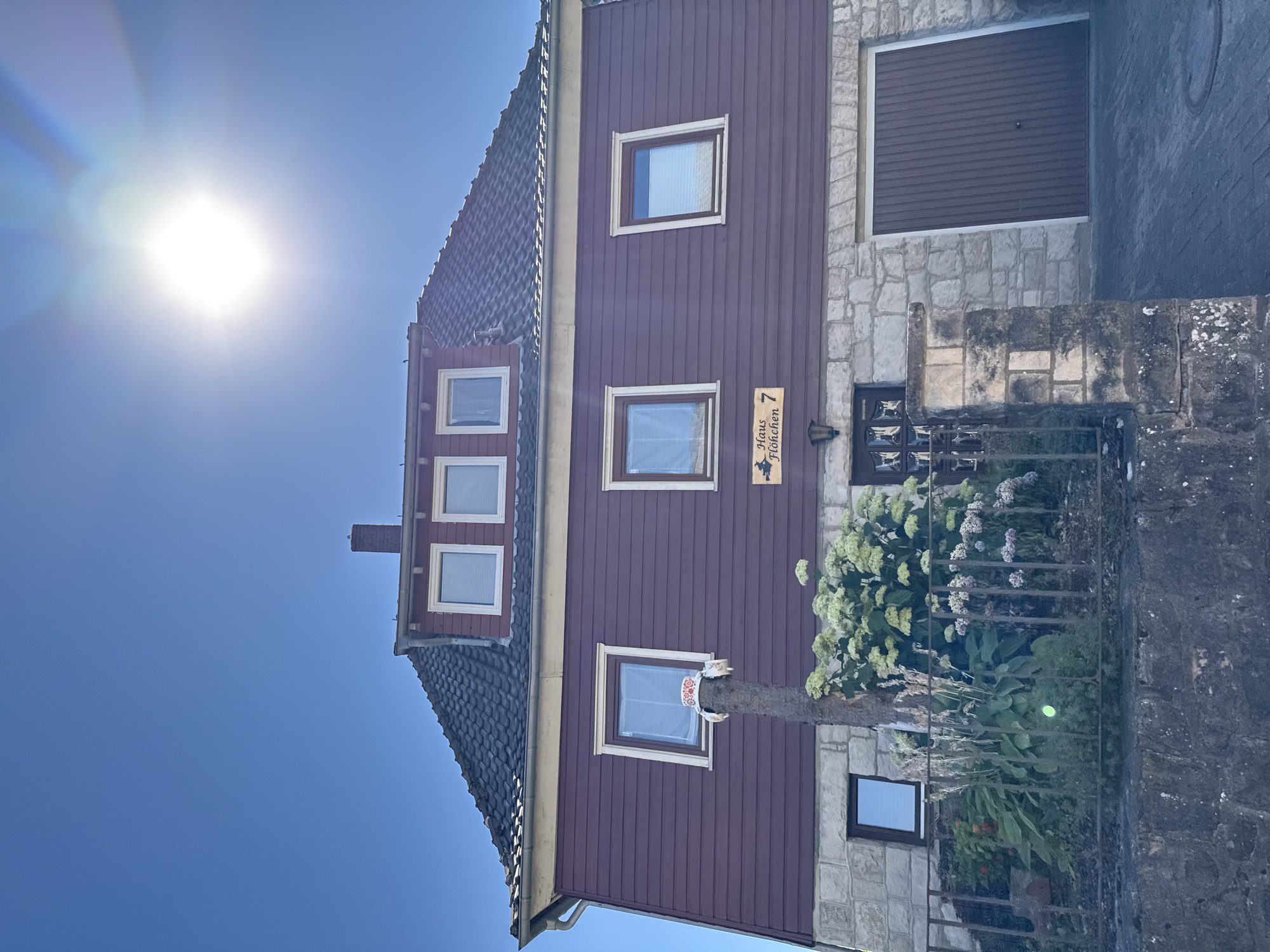 A low-angle, eye-level exterior, full shot captures the front of a two-story house with a dark red wooden exterior on its upper level and a rough stone exterior on its lower level. The house has a dark grey, tiled, pitched roof with a single brick chimney. Three rectangular windows with white trim are visible on the upper level, two of which have white blinds covering them. Two more identical windows are on the middle level of the house, one on the left with a white blind and the other on the right with a clear pane reflecting the sky. Below the right middle-level window is a sign with a black silhouette of a witch on a broomstick to the left and "Haus Flöhchen 7" in black text to the right. The lower level has a dark wood garage door on the right and a dark wood door on the left with a glass pane. A small garden is visible in front of the house, with a black metal fence separating it from the paved driveway. A prominent, tall, bare tree trunk with a decorative element at its top stands in front of the left side of the house, obscuring some of the windows. The sun is visible in the upper right corner of the image, creating lens flare. The sky is a clear, bright blue. The house is located at Haus Flöhchen 7, in Bad Sachsa, Germany.