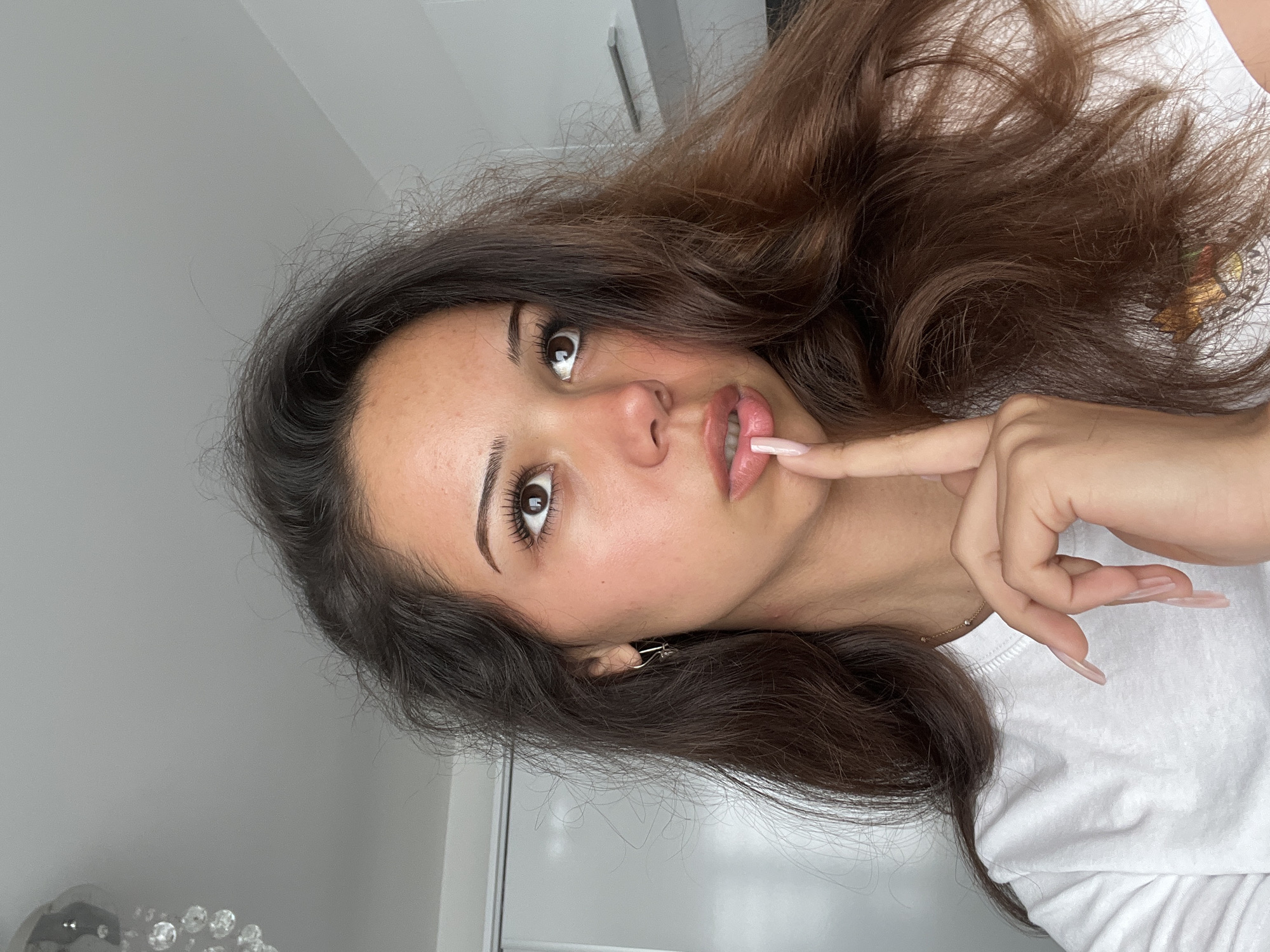 A young woman with long, dark brown hair is shown in a close-up portrait. She is wearing a white t-shirt and has her right index finger touching her lips. Her eyes are looking upwards and to the right, suggesting contemplation or thought. The background is blurred, but appears to be an indoor setting with a white wall and possibly a closet or door on the left side. A chandelier is visible in the upper left corner. The overall impression is of a candid moment captured in a domestic environment. There is no indication of the city of Zlatograd, Bulgaria, or any other specific geographical location.