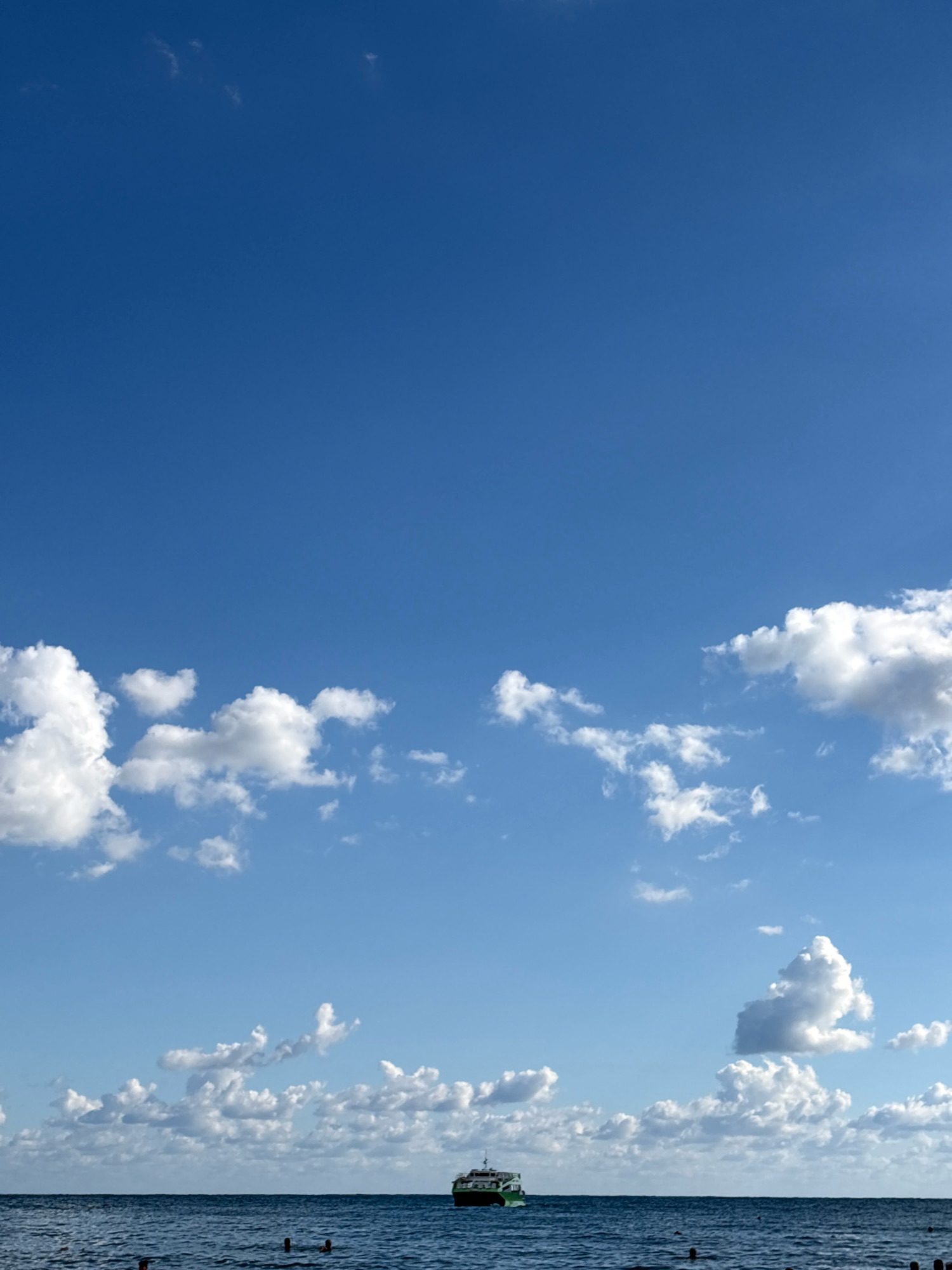 A bright, wide shot shows the tranquil sea meeting a vibrant blue sky scattered with fluffy white clouds. In the foreground, the deep blue ocean is dotted with small, dark posts that appear to be partially submerged. A few swimmers are visible in the water, their heads just above the surface. On the horizon, a green and white boat sails from left to right. The scene suggests a peaceful day at the beach in Arkhipo-Osipovka, Russia.