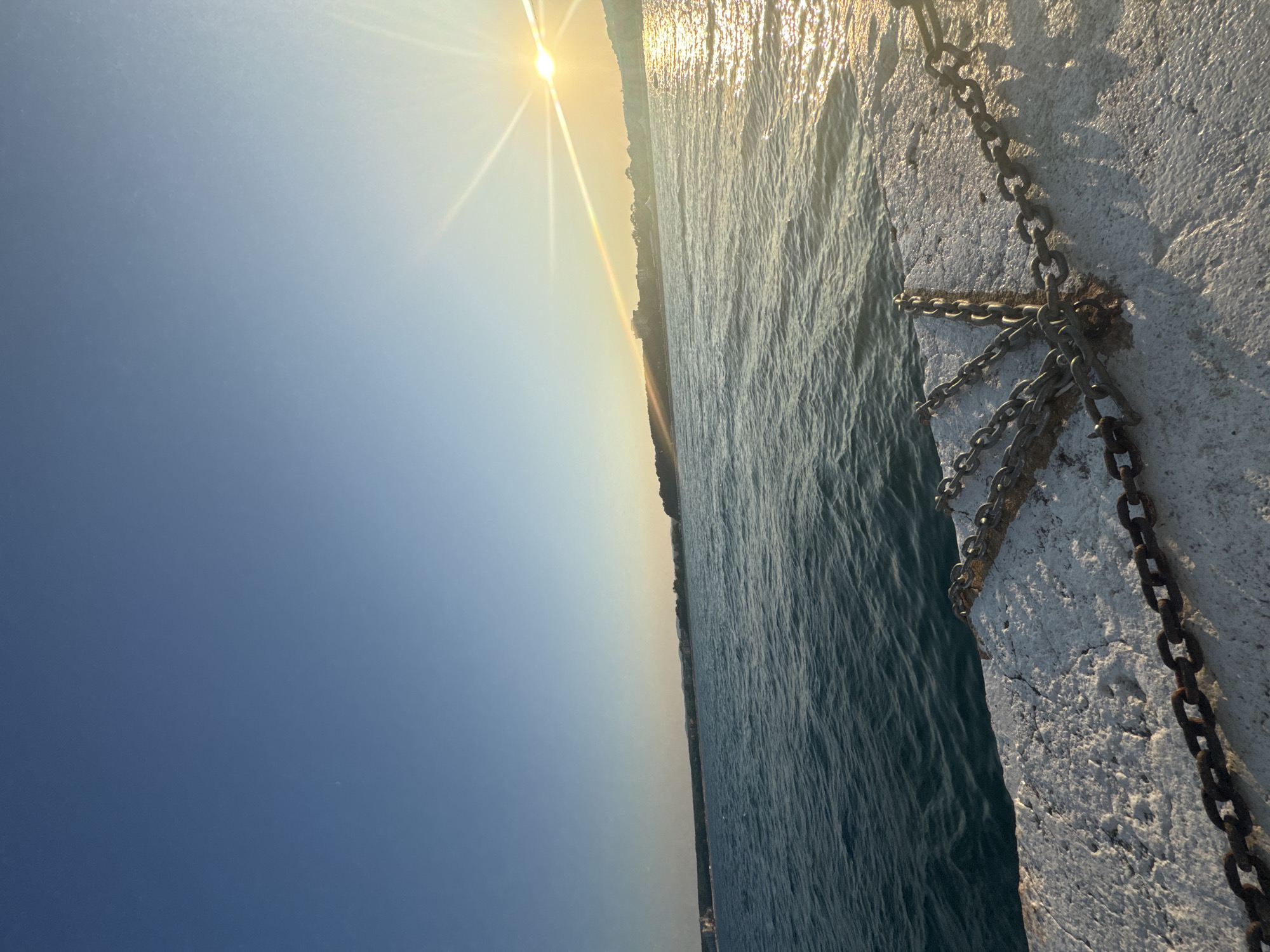 An exterior, eye-level shot captures a sunny, tranquil scene from a pier or breakwater in Primorsko, Bulgaria. The foreground is dominated by a rough, white concrete structure with several thick, dark metal chains draped across it. The chains appear weathered and connect to points out of frame. Beyond the concrete, the deep blue sea stretches to the horizon, with gentle ripples on its surface reflecting the sunlight. In the distance, a long, low-lying coastline is visible, lined with buildings and trees. The sun, a bright orb, is positioned in the upper right quadrant of the image, casting a strong glare and creating shimmering reflections on the water. The sky above is a clear, pale blue, with a gradient transitioning to a warmer yellow-orange hue near the horizon, indicating either sunrise or sunset. There are no people or animals visible in the frame.