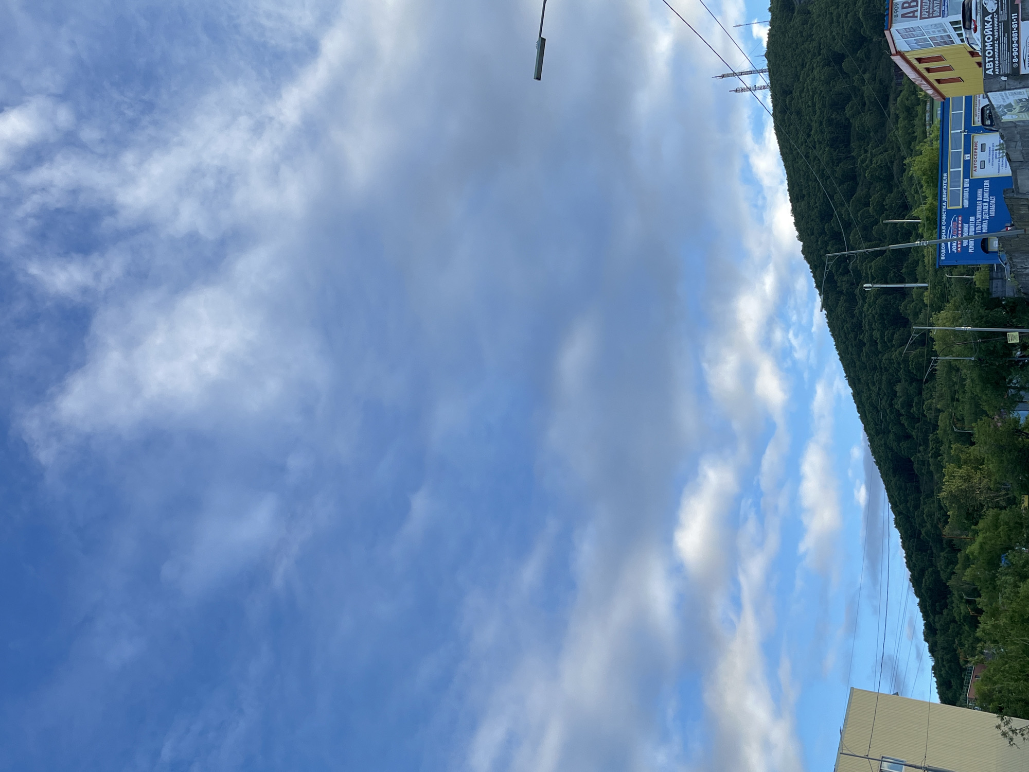The image shows a view of a hillside covered in dense green trees, with buildings and infrastructure visible at its base and on the slope. The sky is a bright blue with scattered white clouds. In the foreground, a portion of a light-colored building is visible on the left. Further to the right, a blue building with white lettering indicating automotive services is present. A yellow building with red accents is located to the right of the blue building. Several streetlights and power lines are also visible throughout the scene. In the distance, radio towers are situated on the hilltop. The overall impression is an urban or semi-urban landscape with natural elements. This scene is consistent with views found in Petropavlovsk-Kamchatsky, Russia, which is known for its mountainous terrain and proximity to natural landscapes.