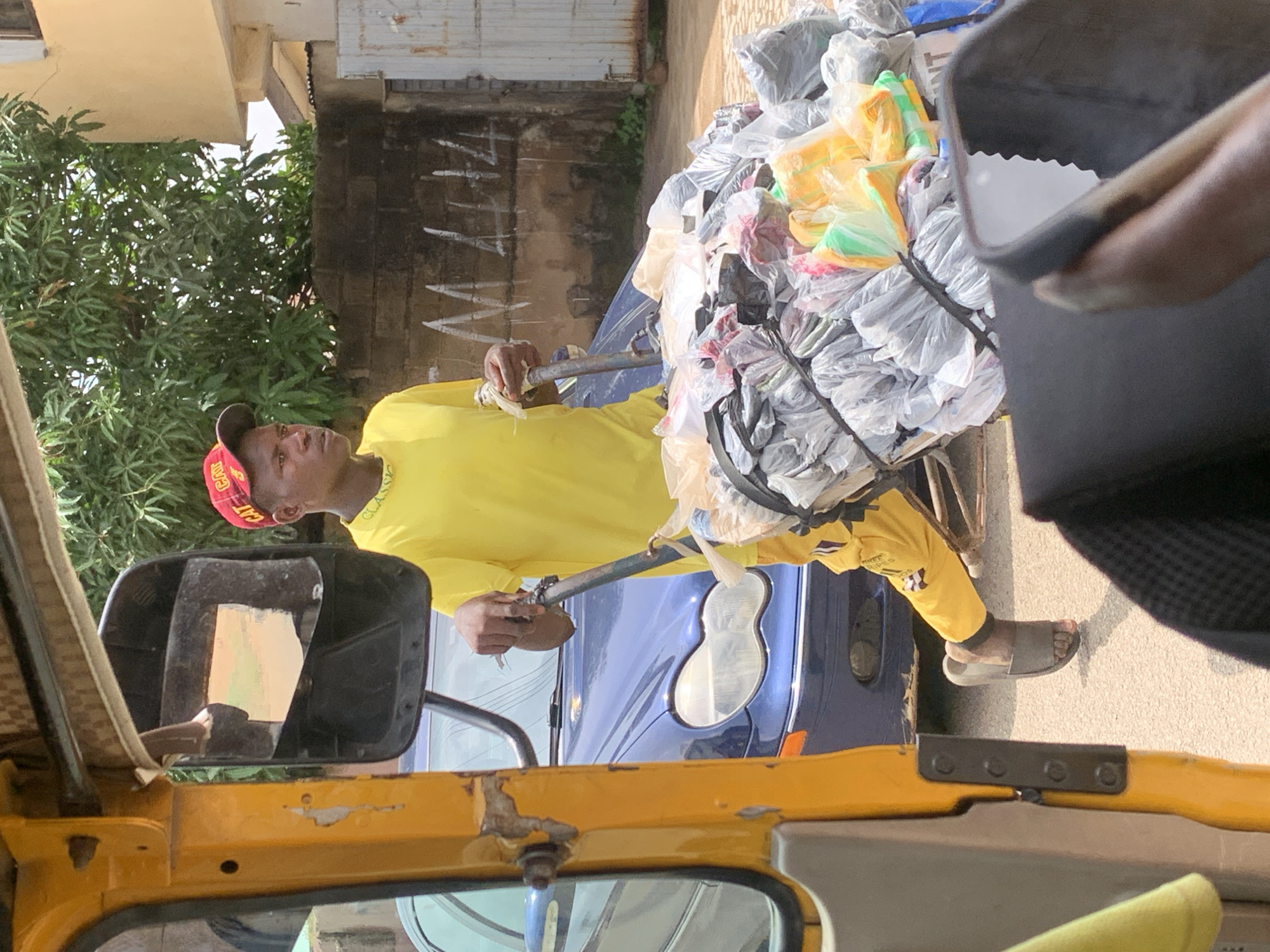 A person wearing a yellow t-shirt, yellow pants with black text, and a red cap is standing next to a pile of plastic bags. The person is holding a metal pole that is attached to the pile of bags. The pile of bags appears to be tied together with black rope. The scene is outdoors, with a building in the background and some greenery on the left. The person is looking towards the left. The ground is a light color, possibly asphalt or concrete. A yellow vehicle, likely a three-wheeled auto-rickshaw, is visible in the foreground, with a side mirror reflecting the scene. The overall impression is that of a person transporting or sorting goods, possibly for recycling or sale. The time of day appears to be daytime, with bright sunlight casting shadows. The weather seems to be fair. Visible text on the red cap reads "CAT". The pants also have some black text, but it is not clearly legible. The context provided indicates the location is Jos, Nigeria.