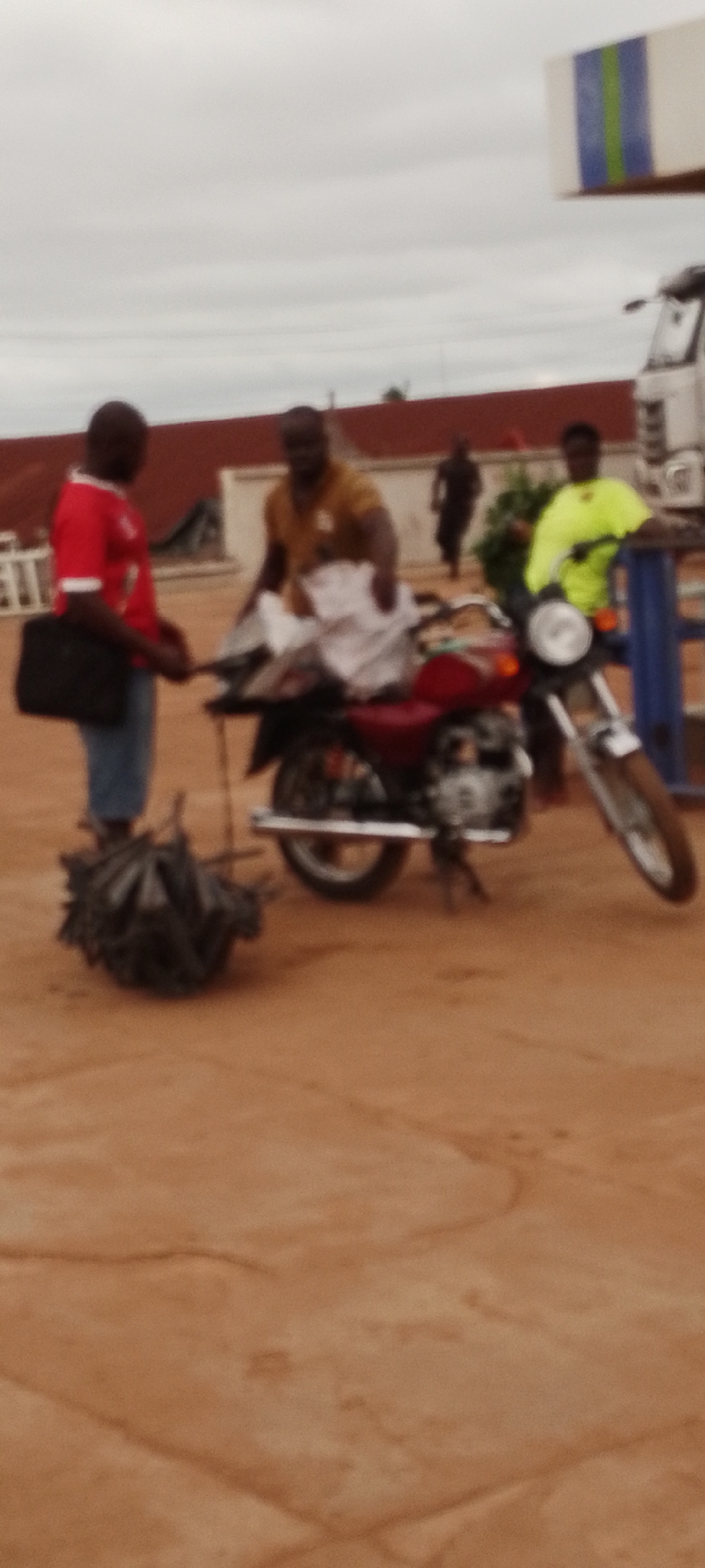 In Gboko, Nigeria, a scene unfolds with several individuals and a motorcycle on a dirt road under an overcast sky. In the foreground to the left, a person wearing a red shirt and blue jeans stands holding a black bag. Beside them, a pile of dark, bundled objects lies on the ground. A motorcycle, predominantly red, is parked in the center, with its kickstand down. A person in a tan shirt is interacting with the motorcycle, appearing to load or unload items, possibly white bags. To the right of the motorcycle, another person is seated on it, wearing a bright green shirt. In the background, another individual is walking away from the camera. The environment appears to be a dusty or sandy area, with a building with a red-tiled roof visible behind the people. A white truck is partially visible to the far right, and a structure with blue and green stripes is positioned in the upper right portion of the image.