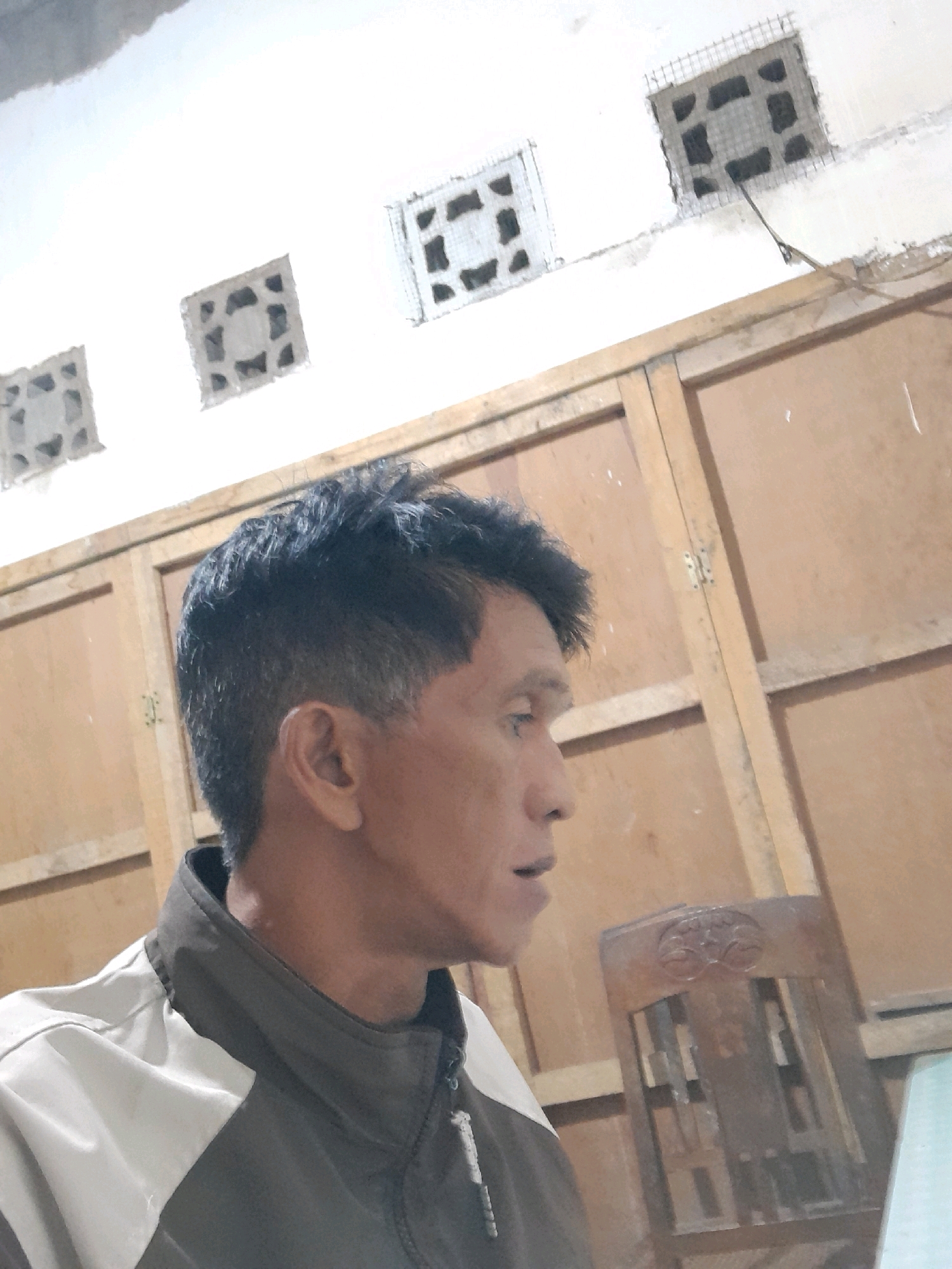 In Glanggang, Indonesia, a man is captured in a medium shot, his face turned to the right, revealing a portion of his neck and shoulder. He is wearing a jacket with a dark gray body and light gray sleeves. The background consists of a wooden cabinet with several square vents set into a white wall above it. The lighting appears to be artificial, casting shadows and creating a somewhat muted color palette. The scene suggests an indoor environment, possibly a residential or utility space.