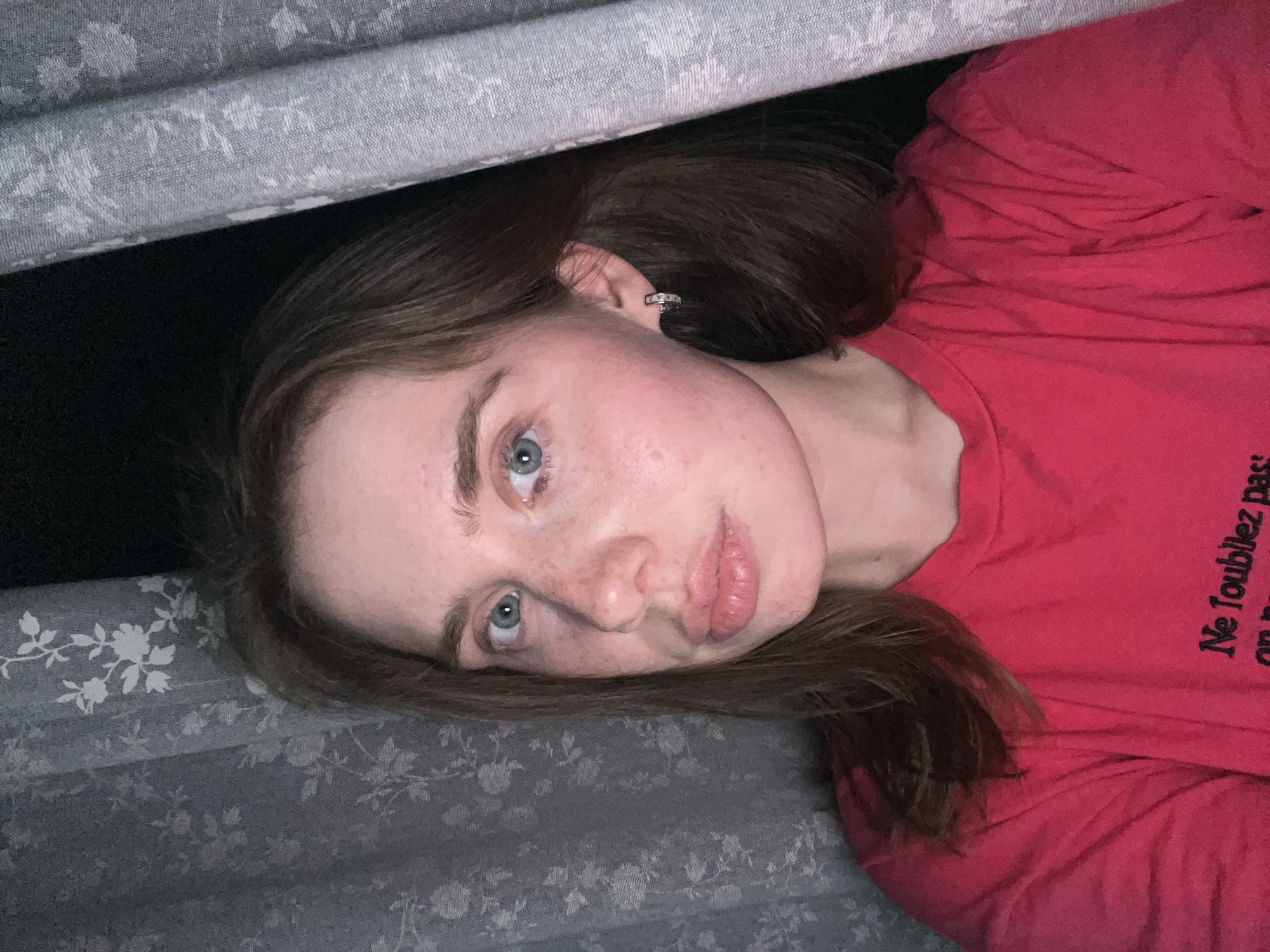 The scene, described as being located in the city of Chaikovsky, Russia, depicts a close-up, chest-up, self-taken portrait of an adult female in a dimly lit indoor environment. The woman has fair skin with visible freckles across her nose and cheeks, blue eyes gazing directly forward, and medium brown, shoulder-length hair. Her lips are full and light pink, slightly parted. She wears a small, silver-colored hoop earring with an etched pattern in her visible left ear. She is dressed in a solid red t-shirt with a crew neck, on which partially visible black text appears to read "Ne Toublez pas" (likely "Ne Troublez pas") on the lower left side. The background consists primarily of two gray or dark blue-gray curtains with a repeating white floral or leafy pattern. The curtain on the left is partially visible behind her, while a more heavily folded curtain on the right borders the frame, obscuring part of her right side. The ambient lighting is low, suggesting a nighttime setting or interior darkness, with a primary light source illuminating the subject's face from the front or slightly above.