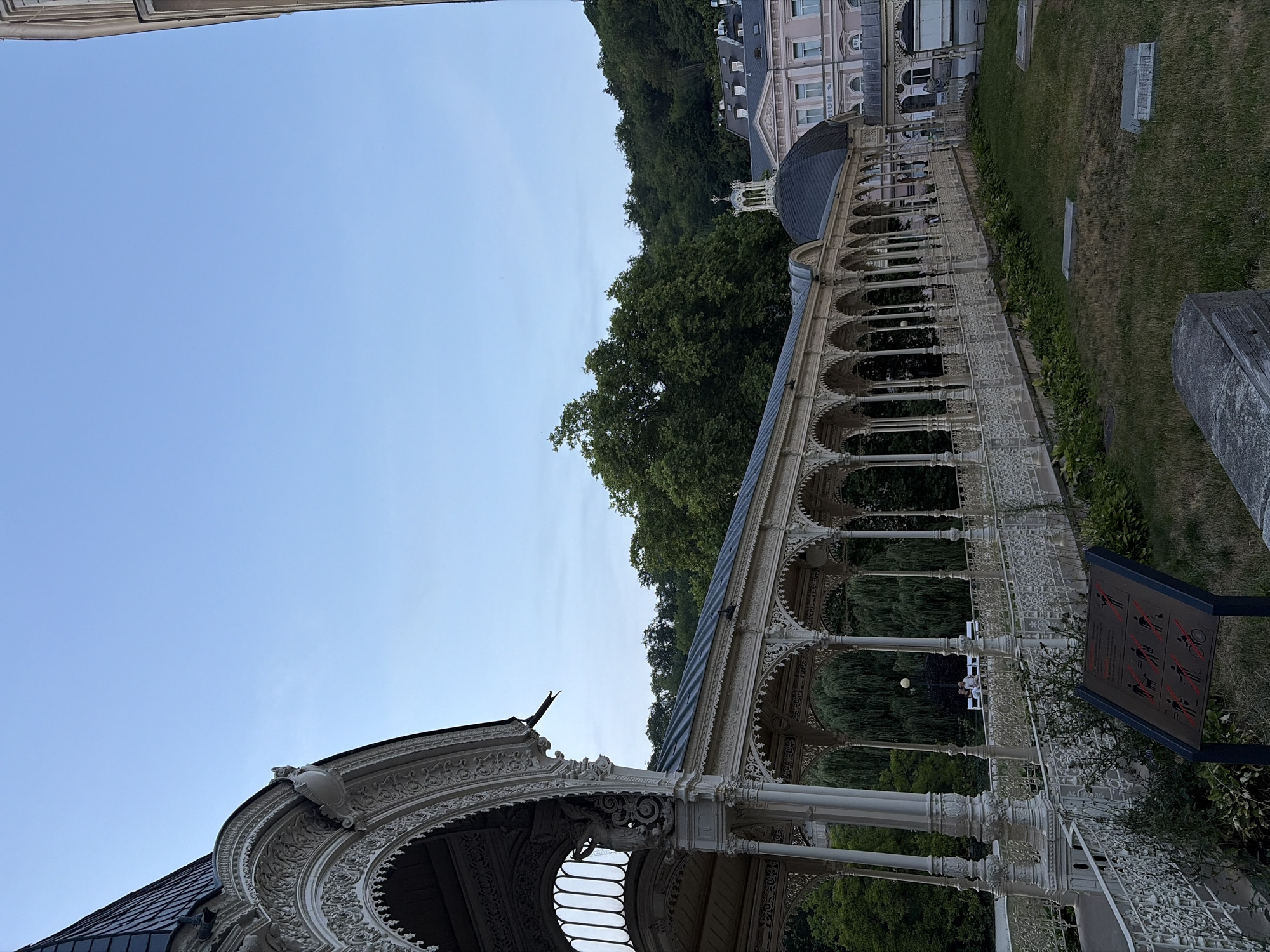 The image shows the Colonnade of the Mill Spring in Karlovy Vary, Czechia, in the late afternoon or early evening. The ornate white cast-iron colonnade stretches across the frame, with its intricate latticework and arched openings. A tiled roof covers the colonnade, and in the background, a large pink building with a domed roof and a smaller cupola can be seen. Lush green trees cover a hill behind the buildings. In the foreground, a grassy area with some scattered stone elements is visible. A sign with red prohibition symbols depicting various activities such as smoking, drinking, and cycling is placed in the lower left corner. Two individuals are seated on a bench in the distance, enjoying the view. The sky is a clear, pale blue.