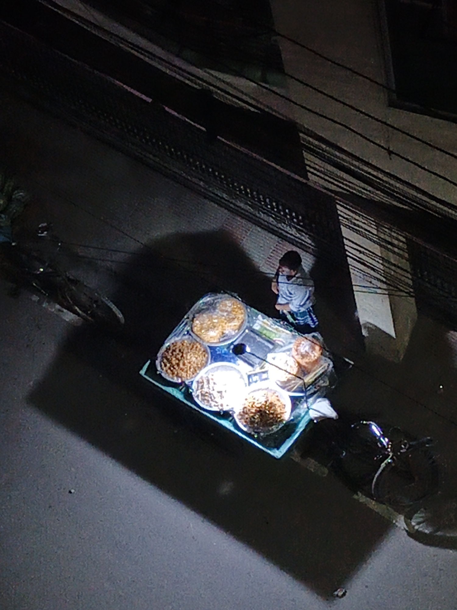 Here's a detailed description of the image:
**Scene:** A nighttime, overhead view of a street scene in an urban environment. The exact location is Unknown, Unknown.
**People:**
* One person is visible standing behind a food cart. The individual is wearing a light-colored shirt and a checkered garment around the waist. The person's stance suggests they are tending to the food cart.
**Objects:**
* **Food cart:** The central object in the image is a brightly lit food cart. It's topped with multiple bowls or containers, presumably containing food items. Some are covered.
* **Bowls/Containers:** Several bowls are displayed with varying contents; likely snacks, desserts, or ingredients.
* **Bicycle:** A bicycle is parked on the left side of the cart.
* **Motorcycle/Scooter:** A motorcycle or scooter is partially visible on the right side of the cart.
* **Overhead cables:** A dense network of overhead cables and wires dominates the upper portion of the image, spanning across buildings.
* **Buildings:** Parts of buildings can be seen in the background.
* **Pavement:** The surface is a dark pavement street.
**Actions:**
* The primary action is a person running a food stall on the street.
**Interactions:**
* The individual behind the cart seems to be interacting with the food on display, presumably preparing or selling it.
**Environment:**
* **Lighting:** It is nighttime, with light emanating from the food cart.
* **Urban Setting:** The presence of buildings, cables, and motorized vehicles indicate an urban environment.