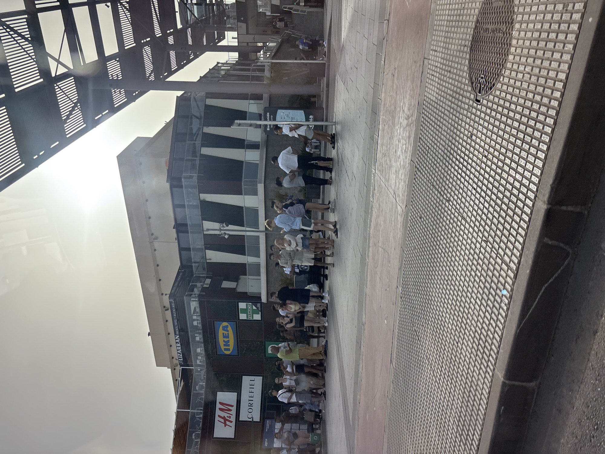 The image shows a street scene in front of a commercial building, likely a shopping center, in Orihuela, Spain. The building features prominent signage for various businesses, including "LIZARRAN" with text that appears to be "RINCONES TAPAS Y MUCHO MAS", "H&M", "CORTEFIEL", "IKEA", and "FARMACIA". A group of people are standing in a line on the paved area in front of the building, some are looking at their phones, others are talking. There is a man in a lime green shirt holding a phone, appearing to be talking to someone. Another man in a white shirt and dark pants is standing in the line. To the right, a metal structure with parallel slats forms a canopy or roof over a walkway or plaza. The ground in the foreground is a textured, checkered metal grate. Sunlight streams in from the upper right, casting shadows and creating a bright sky.