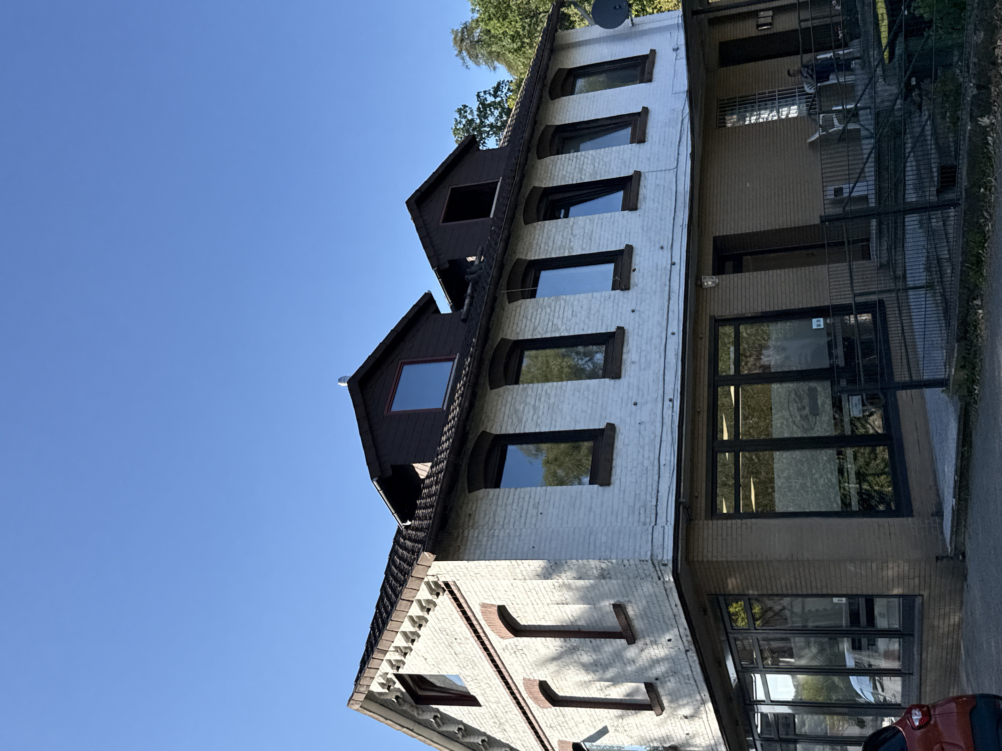 The image shows a building located in Osterode am Harz, Germany. The building has two stories above a ground-level commercial space with large windows. The upper story is white brick with dark-trimmed windows, some of which are arched. The roof is dark with dormer windows. The ground level features brown brick and expansive glass storefront windows. A black metal fence encloses an outdoor seating area on the right side of the building, where a person is seated in a chair. A red car is partially visible in the lower left corner of the frame. The sky is clear and blue.