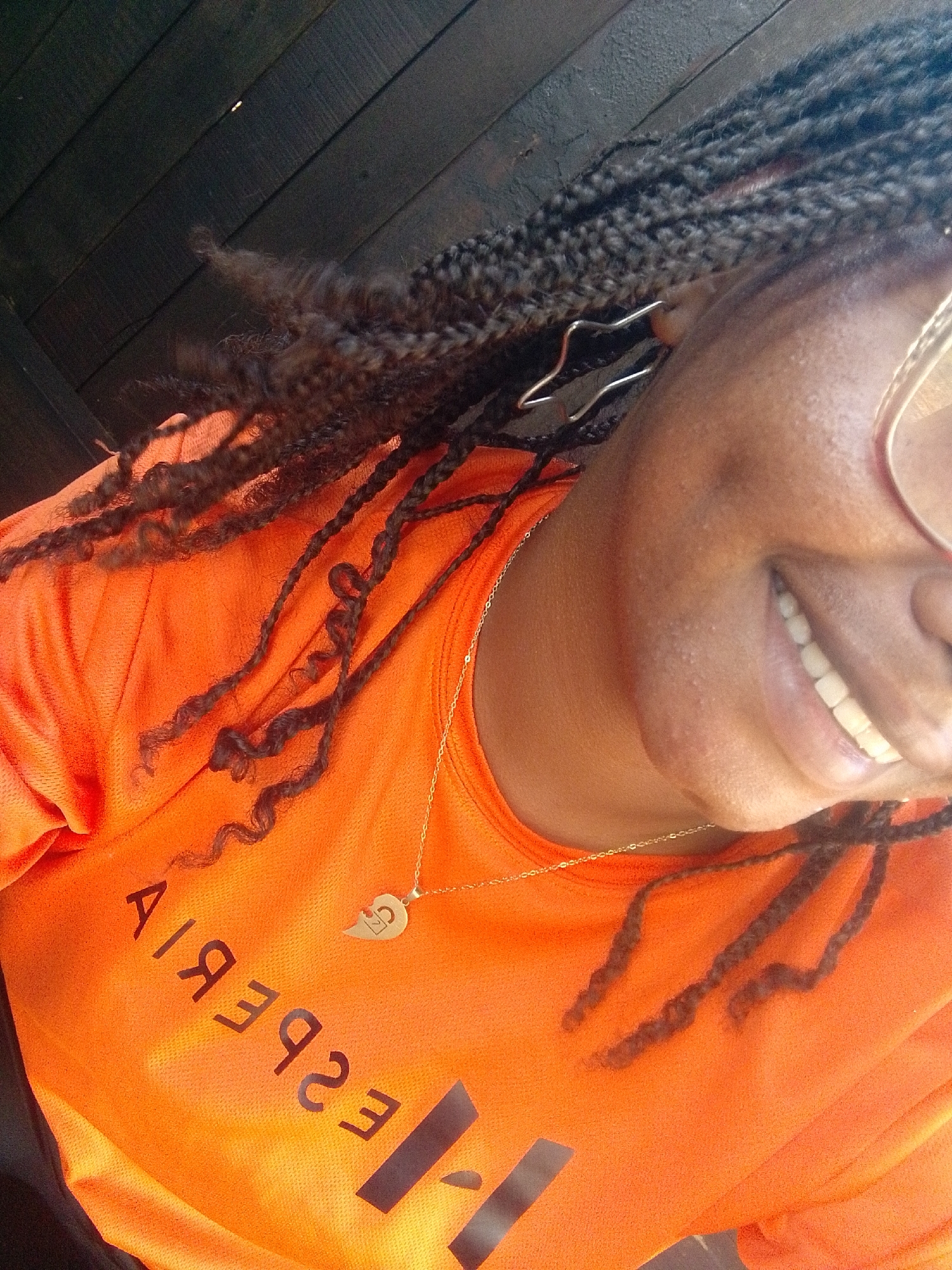 This angled, close-up portrait, taken in Pedro González, Venezuela, features a person with dark skin and long, dark braided hair, smiling and looking upwards. The subject wears a bright orange short-sleeved shirt with reversed text, which reads "ESPERIA" and a partially visible, reversed dark graphic, possibly a large "H". A delicate gold chain necklace with a small, gold heart-shaped pendant, appearing to be a split heart design with the initials "LC" on one half, rests on the shirt. The person also wears gold-rimmed glasses, partially visible over the right eye, and a unique silver-toned, abstract metal earring in the left ear. The background consists of dark wooden planks, suggesting an overhead ceiling or structure, possibly in an indoor or sheltered outdoor setting. The bright lighting indicates it is daytime. The clear smile on the subject's face conveys happiness or contentment.