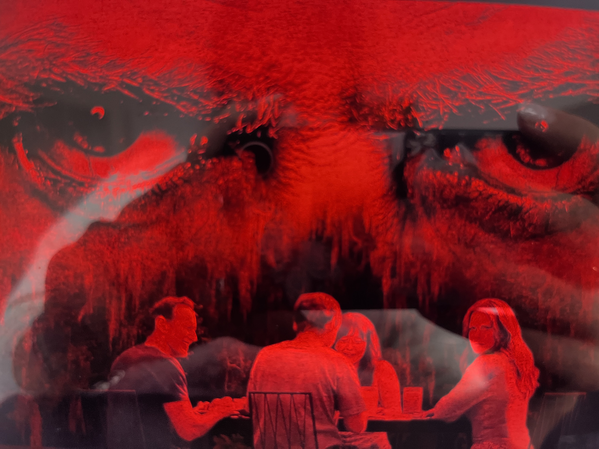 The image shows four people seated at a table under a large, abstract red form that resembles a stylized face or creature. The environment is dimly lit, with the dominant color being red, suggesting artificial lighting or a filter. The red form in the background appears to be projected or a structural element of the location.
On the left, a man with short dark hair is seated facing right, looking towards the table. He is wearing a dark t-shirt. To his right, another man with short dark hair is seated facing the table, wearing a lighter colored t-shirt. He is partially obscured by a chair. In the center, a person's head and shoulders are visible, facing the table. To the right of the center, a woman with long, curly dark hair is seated facing left, looking towards the viewer and over her shoulder. She is wearing a patterned garment.
The table in front of them is dark and appears to have drinks on it. The overall impression is a dimly lit indoor scene with a dramatic and somewhat surreal background. There is no discernible information in the image that directly indicates the location in the city of Gertwiller, France.