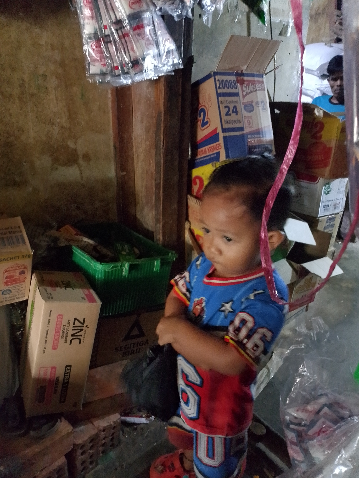 A young child, approximately 2-3 years old, stands in what appears to be a market or a small shop. The child is wearing a blue t-shirt with white stars and red and white striped sleeves and shorts with the number "06" prominently displayed. The child's head is turned to the left, and their expression is neutral.
The background is cluttered with various goods, suggesting a commercial setting. There are stacks of boxes, some labeled with Indonesian text like "Sukses" and "ZINC ANTI DANDRUFF SHAMPOO," indicating the sale of consumer products. A green plastic container is filled with items, possibly more merchandise. Hanging from the ceiling are several clear plastic packages containing small, white-wrapped items. A bright pink ribbon or plastic strip hangs down prominently in the foreground, partially obscuring the child. In the far background, a person wearing a blue shirt is visible.
The overall environment is dimly lit, with a rough, textured wall on the left side of the image. The floor appears to be made of concrete or similar material. The presence of goods commonly found in Indonesian markets and the style of packaging suggest this scene could be located in Palembang, Indonesia.