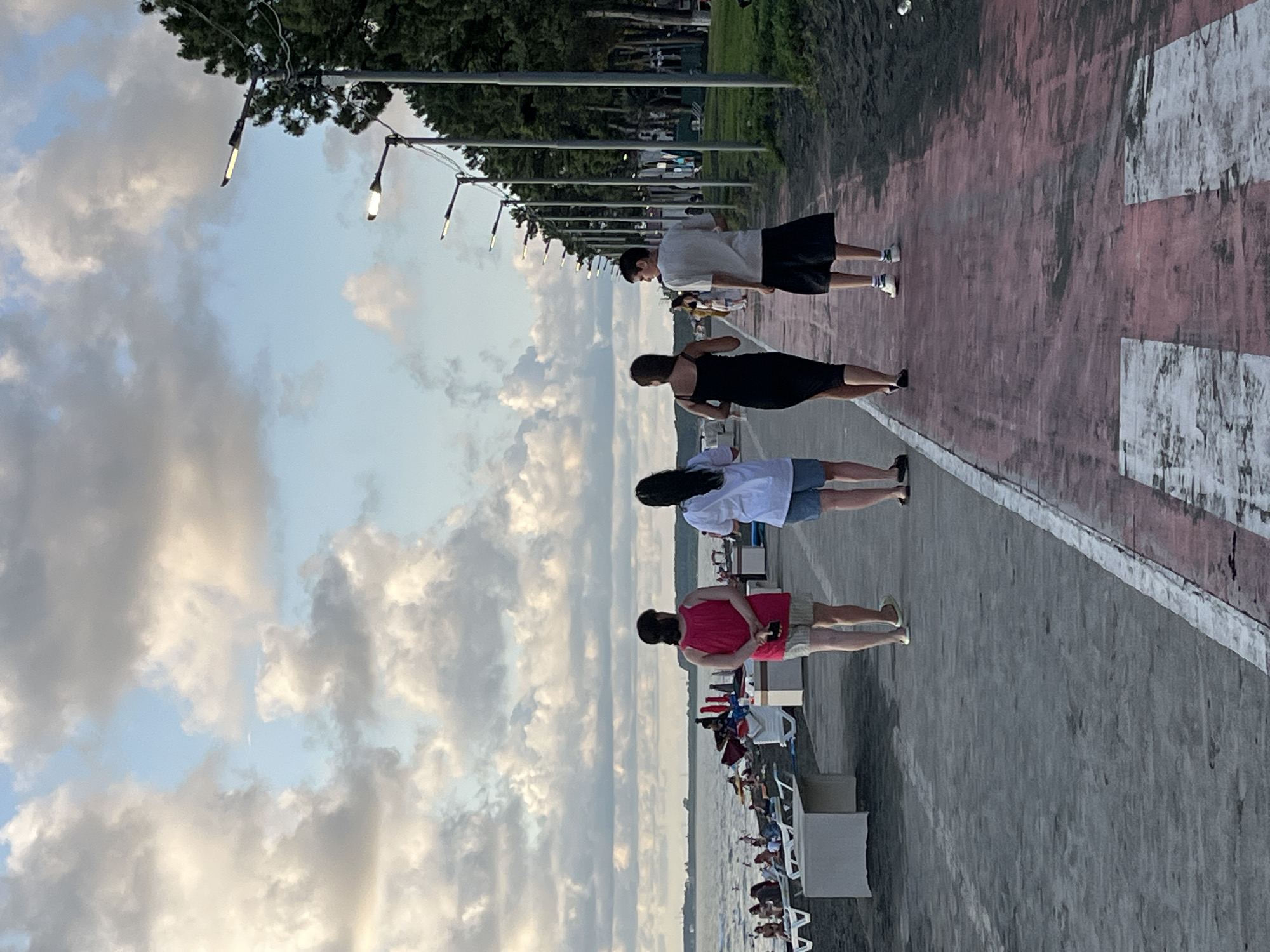 A group of four individuals, two women and two men, are walking away from the camera on a paved walkway next to a black sand beach in Ureki, Georgia. The walkway has a red-colored asphalt section with white painted lines, resembling a crosswalk. The beach is lined with lounge chairs and umbrellas. In the background, the Black Sea is visible under a cloudy sky. On the right side of the walkway, a row of modern streetlights and tall green trees line the path. The group appears to be casually strolling, with the two women in front and the two men behind them.