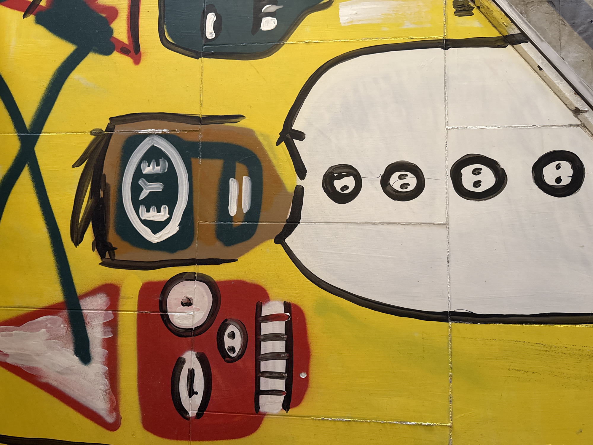 The image shows a close-up section of a colorful, cartoon-style mural painted on a yellow surface, likely a wall. The mural features several abstract and stylized figures. On the left, there's a brown shape containing a white oval with the word "EYE" written vertically inside. There's a red square with a white circle, a white oval, and a series of white vertical lines along the right edge. To the right of the brown shape is a large white shape with four black circles, each containing two white dots. A green diagonal line crosses part of the image. The artwork is clearly painted with thick lines and bold colors. The surface has visible lines suggesting it's divided into sections, possibly tiles or panels. There is no evidence of the city Moscow, Russia or any people, interactions or actions occurring.