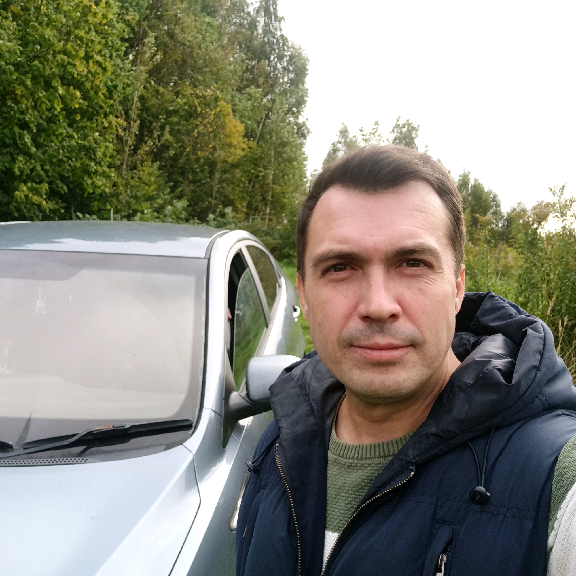 A middle-aged man with short, dark hair and a neutral expression is taking a selfie outdoors next to a light-colored car. He is wearing a dark blue hooded vest over a green knit sweater. The car's driver's side door is slightly ajar, and a person can be seen in the driver's seat. The background consists of green trees and foliage, with a light sky visible above. The scene appears to be in a rural or park-like setting, possibly on the outskirts of Nizhny Novgorod, Russia, given the presence of mature trees and a less urbanized environment.