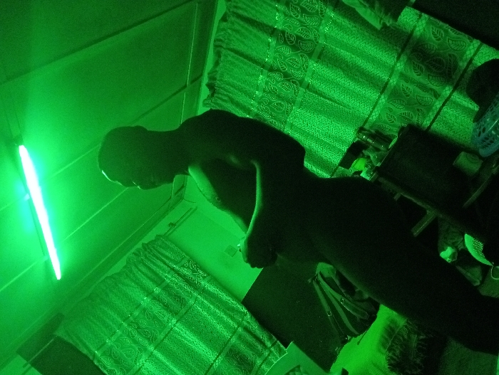 A person stands silhouetted in a room lit with green light. The light source, a fluorescent tube, is visible on the left wall. Sheer curtains hang on two walls, and a table with various items is partially visible on the right. The perspective is skewed, looking up at the person and the room's contents.
