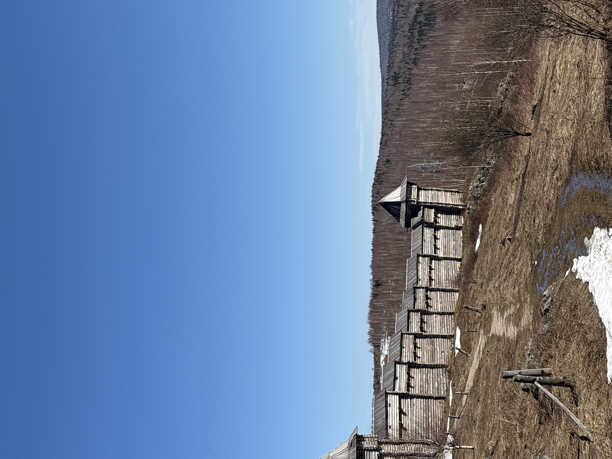 The image depicts a wooden structure, possibly a fortified wall or tower, located near the city of Gubakha, Russia. The structure is constructed of logs and consists of multiple tiers, with a prominent tower element at one end. Adjacent to the structure is a landscape of dry vegetation and remnants of snow, suggesting early spring. Beyond the structure, a dense forest stretches along the horizon. The sky is a clear blue. The location is identifiable as Gubakha due to the distinct architectural style of the wooden structure, characteristic of historical fortifications in that region.