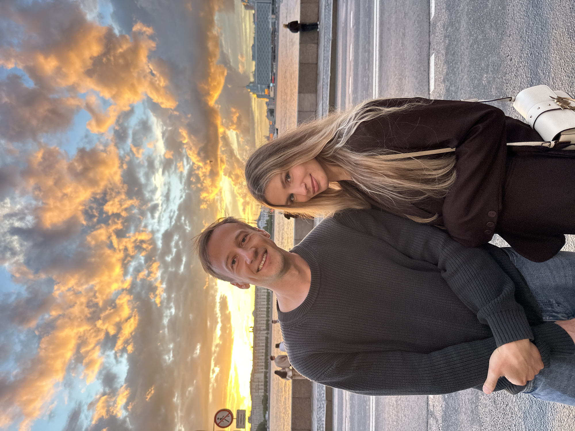 The image shows a young Caucasian couple standing on a city street next to a river during sunset. The sky is filled with dramatic orange and yellow clouds. In the background, across the river, is a cityscape with buildings, including what appears to be a hotel with illuminated windows. A pedestrian walkway with a railing separates the street from the river. On the left side of the image, a sign with a red circle and a line through it is visible, indicating a prohibition. Several other people are seen in the background, walking along the riverfront. The man in the foreground is wearing a dark gray sweater and blue jeans, and the woman is wearing a dark brown jacket and carrying a white handbag. They are both looking at the camera and smiling. The overall impression is of a romantic evening in Saint Petersburg, Russia.